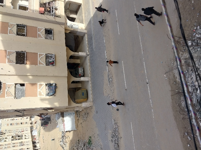 An elevated view captures an urban street scene in Khan Yunis, Palestinian Territory. In the midground, an asphalt road with faded white lane markings spans horizontally. Six individuals are visible on the road: one male youth in dark clothing on the left is dribbling a light-colored football; another younger male in an orange-brown shirt walks right, away from the first youth. To the right, two adult males walk towards the right; further right and partially obscured, two smaller figures are in motion, one crouched low, the other running.
Behind the road, a multi-story building (at least four visible floors) with a beige facade and light brown vertical panels dominates the right-center. This building displays significant damage, including broken windowpanes, missing glass, and potentially impact marks on its exterior. Several windows feature decorative arched elements. The ground floor incorporates arched openings, sheltering a light-colored sedan and an olive-green, possibly covered, vehicle.
To the left of this primary building, an open, dusty area is strewn with rubble and debris. A small, white, tent-like structure stands in this area. Further back on the left, another multi-story building, also appearing damaged, is visible. Long shadows extend across the open ground.
In the immediate foreground, at the bottom of the image, multiple power lines or cables with black and white insulation are stretched horizontally across the frame, obscuring part of the ground which consists of rubble and sparse vegetation.