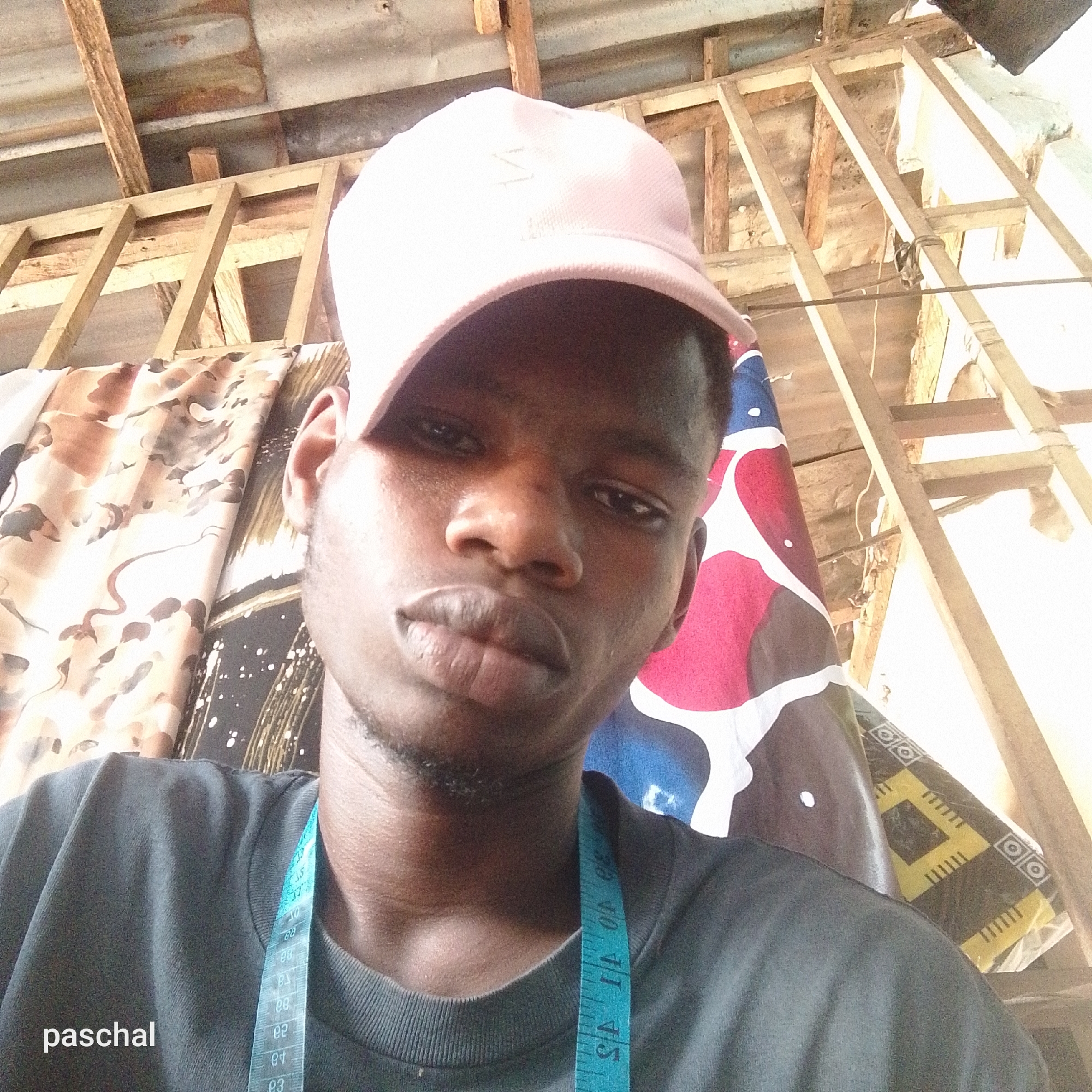 A close-up selfie of a young Black man with short hair and a light pink baseball cap, possibly inside a building in Nsugpe, Nigeria. He is wearing a gray shirt and a blue measuring tape is draped around his neck. Behind him are patterned fabrics and a wooden frame construction. The ceiling appears to be made of corrugated metal. The name "paschal" is visible at the bottom left of the image.