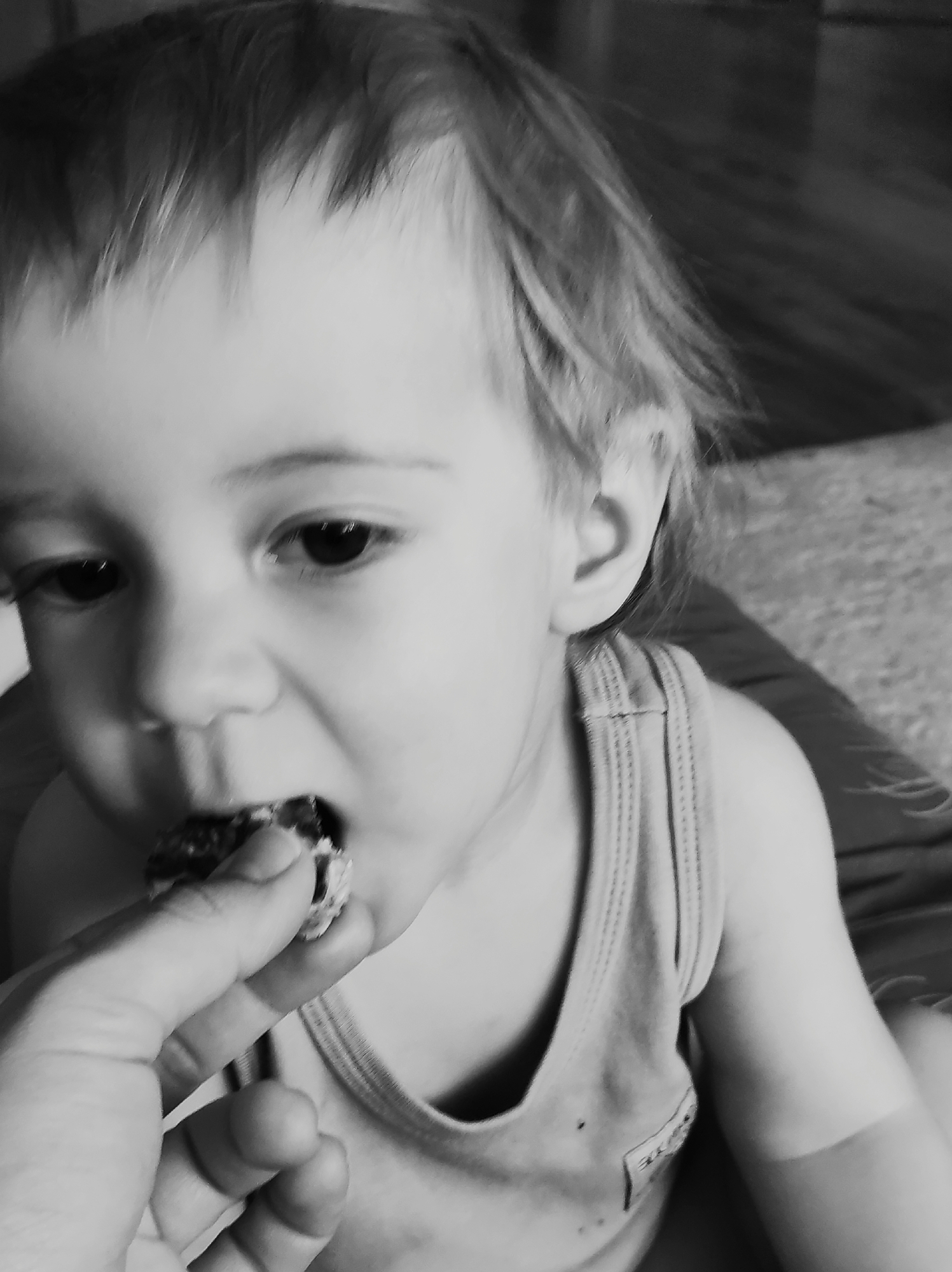 A monochrome, close-up shot depicts a young child being fed. The child, fair-skinned with light hair, is wearing a tank top and is in the process of eating something being offered. A hand, presumably belonging to an adult, is holding a small portion of food close to the child's mouth. The child's gaze is directed downwards towards the food. The background is blurred and indistinct. There is no way to discern the location is in Chaikovsky, Russia.