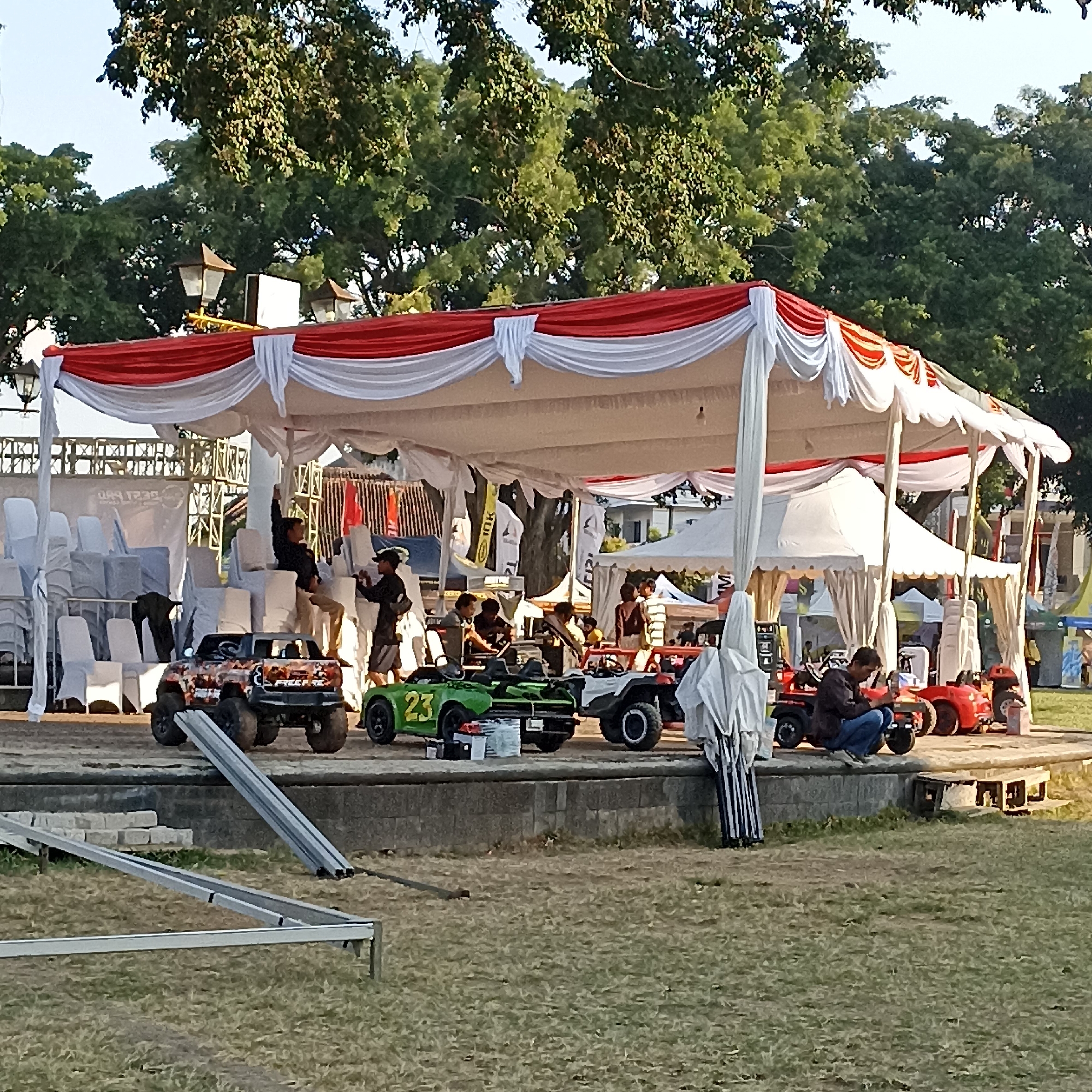 The image shows an outdoor event in Magelang, Indonesia, likely at a park or public space, given the presence of trees and grassy areas. A large canopy tent decorated with red and white fabric serves as the main structure. Underneath the tent, several children's ride-on toy cars are displayed, including a green one with the number "23" on it, and a more rugged, colorful car with large wheels. People are scattered around the area, some appearing to be supervisors or organizers. One man is sitting on a red toy car, looking at his phone. Another person is standing by the green car, possibly attending to it. In the background, stacks of white chairs are visible, suggesting seating for an audience or participants. The overall atmosphere appears to be casual and perhaps family-oriented, with the toys indicating an activity for children.