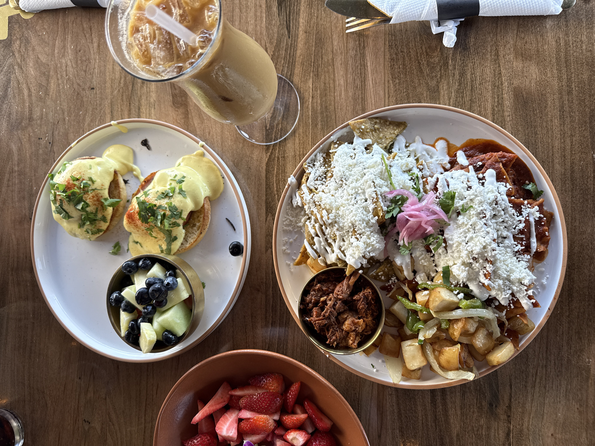 A high-angle shot shows a wooden table set with food, likely in a restaurant in Chula Vista, California, United States of America. On the left, there is a white plate with two servings of what appears to be eggs benedict, accompanied by a small bowl containing blueberries and melon. Next to the plate is a tall glass filled with iced coffee or a similar beverage. To the right, another white plate displays a portion of chilaquiles, a dish of fried tortilla chips in sauce, topped with cheese and possibly other ingredients. A small metal bowl filled with a dark, shredded meat sits next to the chilaquiles, and a bowl of cut strawberries is placed near the bottom of the image. A knife and fork wrapped in a white napkin are placed at the top right. The table exhibits a warm, natural wood grain, and the overall presentation suggests a brunch or lunch setting.