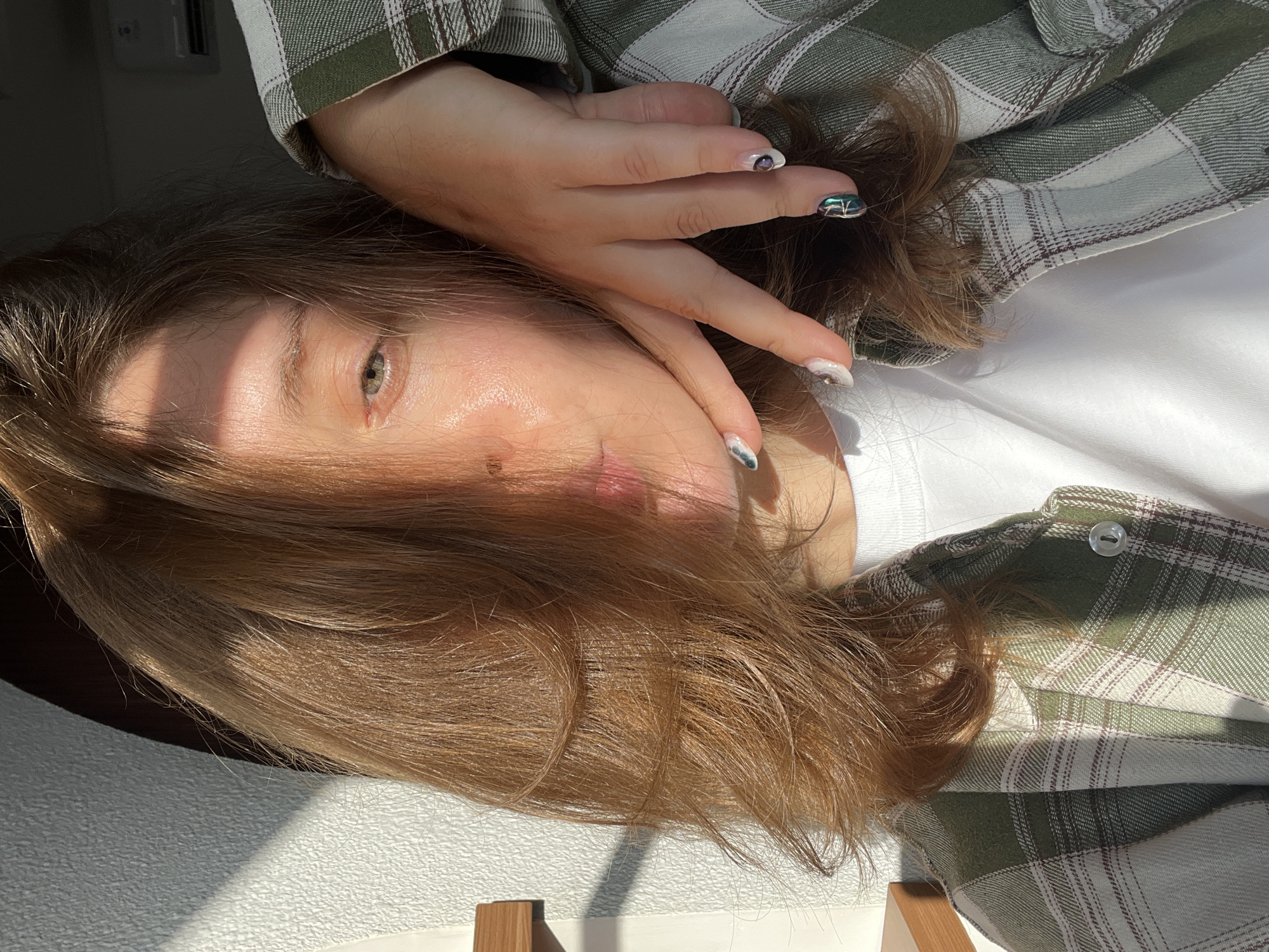 A person, likely female, is lying down, facing the camera. She is wearing a white t-shirt and a green and white plaid shirt. Her right hand is raised, with her fingers touching her cheek. She has shoulder-length brown hair. The light is strong, casting shadows across her face. The location cannot be determined from the image and no association can be made with North Kuta, Indonesia.