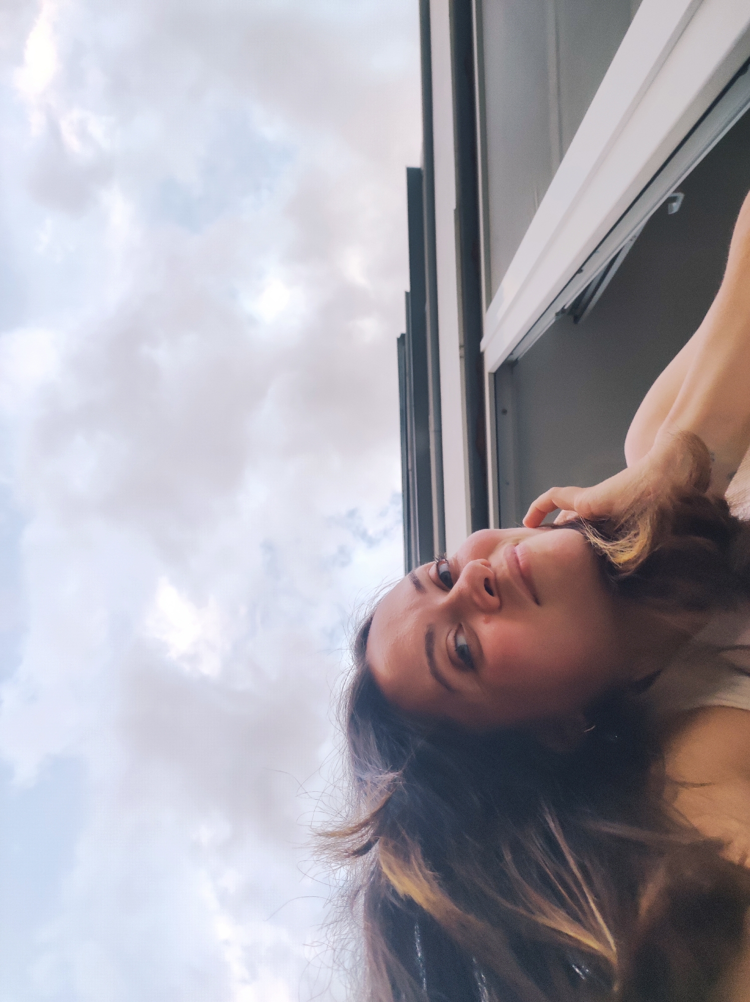 A young woman is positioned at an angle, looking up and to the left towards the camera. Her face is partially obscured by her dark hair, which has some blonde highlights. She has a hand raised near her face. The background shows a sky filled with cloudy formations. A window frame with a dark grey exterior and white trim is visible, partially framing the scene. It is impossible to determine the location in the city of Chaikovsky, Russia, based on this image.