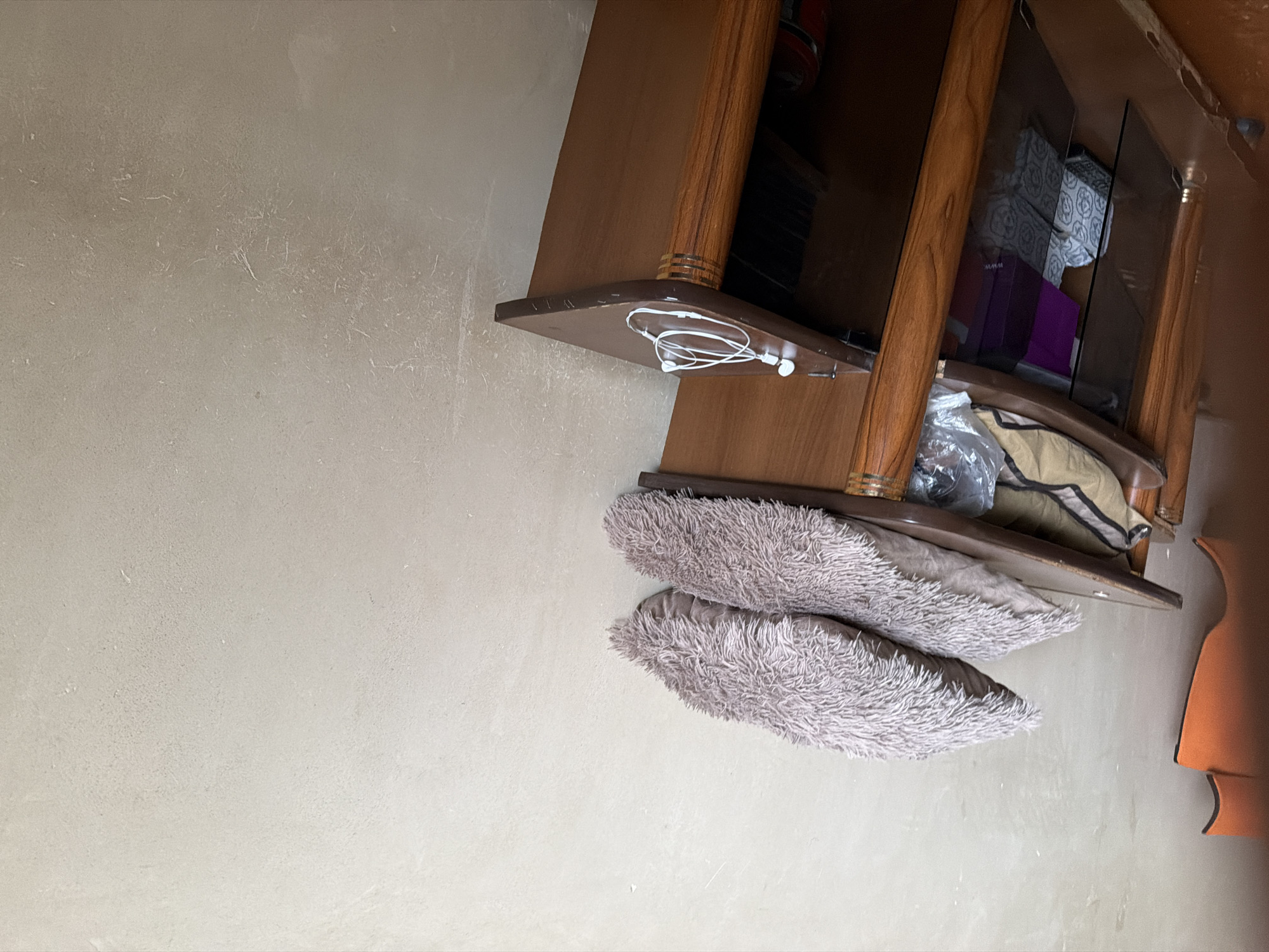 In Tandzut, Armenia, an indoor scene shows a wooden entertainment unit against a textured, beige wall. The top shelf of the unit holds two fluffy, light brown pillows stacked on top of each other. Below, another shelf contains various items including a tan and green bag, a clear plastic-wrapped object, and several decorative boxes, one purple and two with a grey and white pattern. The lower part of the unit features glass doors, revealing further storage. On the surface of a lower shelf, a pair of white earbuds are casually placed. The overall impression is one of a domestic setting with items suggesting recent use or ongoing organization.