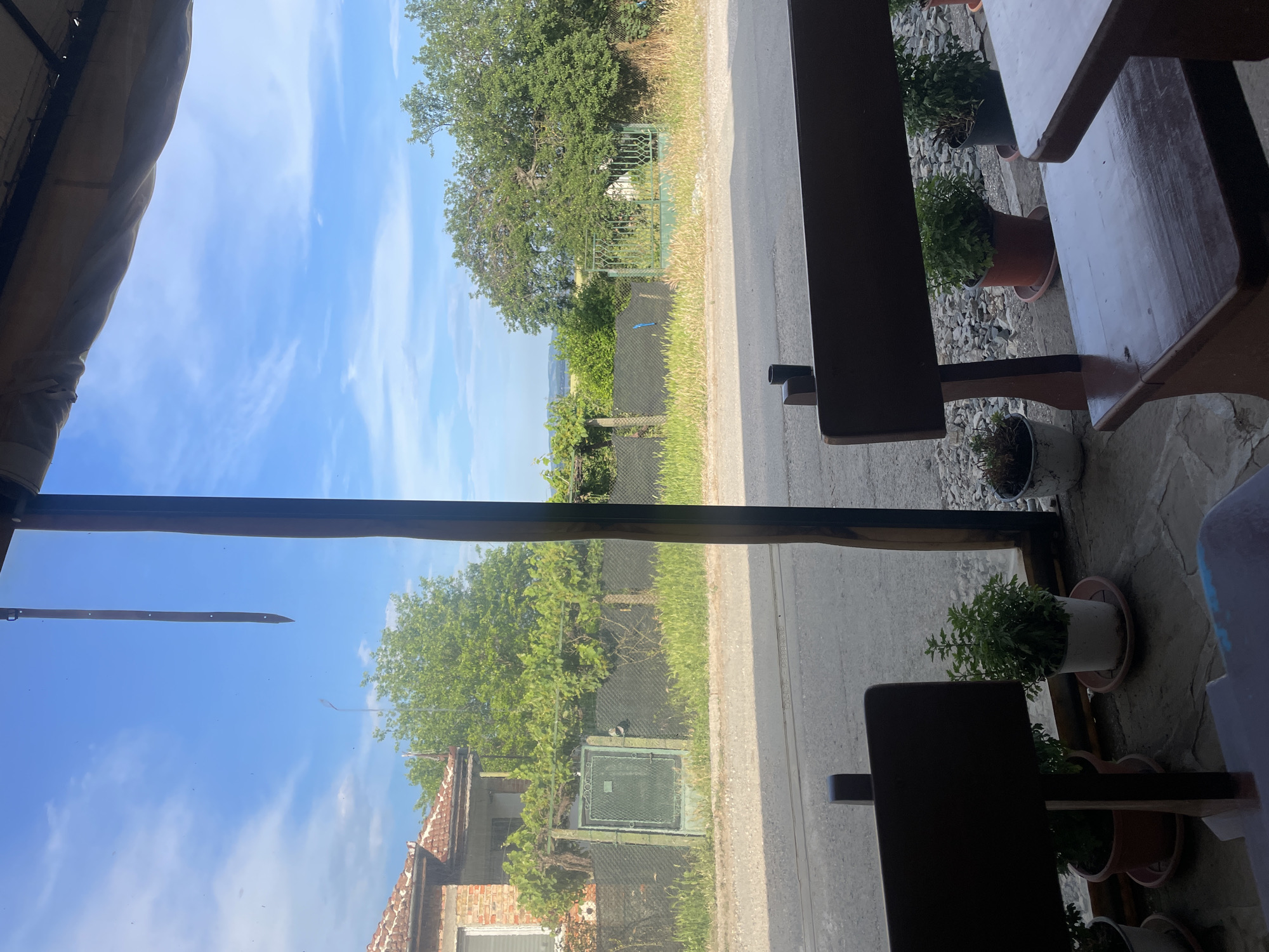 This image presents a static, daytime view looking out from an enclosed or semi-enclosed structure in Konstantinovo, Bulgaria, towards a rural or semi-rural street scene.
In the immediate foreground, dark wooden benches are partially visible, positioned with their backs towards the viewer, suggesting an outdoor seating area. On the right, a dark wooden table is also partially seen. Several potted plants, some in terracotta and some in white or black pots, are arranged on a flagstone or paved floor directly behind a transparent barrier. A dark vertical wooden or metal post, part of the framing for the transparent barrier, is prominent on the mid-right, with another partial post on the far left. A dark, narrow strip of material hangs vertically from the top edge of the frame, slightly left of center, against the sky. An awning or roof structure casts a shadow at the very top of the frame.
Through the transparent barrier, a paved road or street stretches horizontally across the mid-ground. Along the far side of the road, a strip of dry grass and weeds is visible. Beyond this, a dark mesh or chain-link fence runs parallel to the road. Two distinct gates are visible along this fence line: a dark green, rectangular metal gate with a solid panel on the left, and a more ornate, lighter green metal gate with decorative scrolling on the right, further down the road. Green foliage, including what appears to be trellised grapevines on the left, grows along and behind the fence.
In the background, on the far left, a brick house with a reddish-tiled roof is situated behind the fence. A white-framed window is partially visible on the house. To the right, beyond the fence, a dense collection of green trees forms a natural backdrop. In the distant background, rolling hills or fields are visible under a bright blue sky marked by scattered white, wispy clouds. No individuals are present, and no actions are depicted.