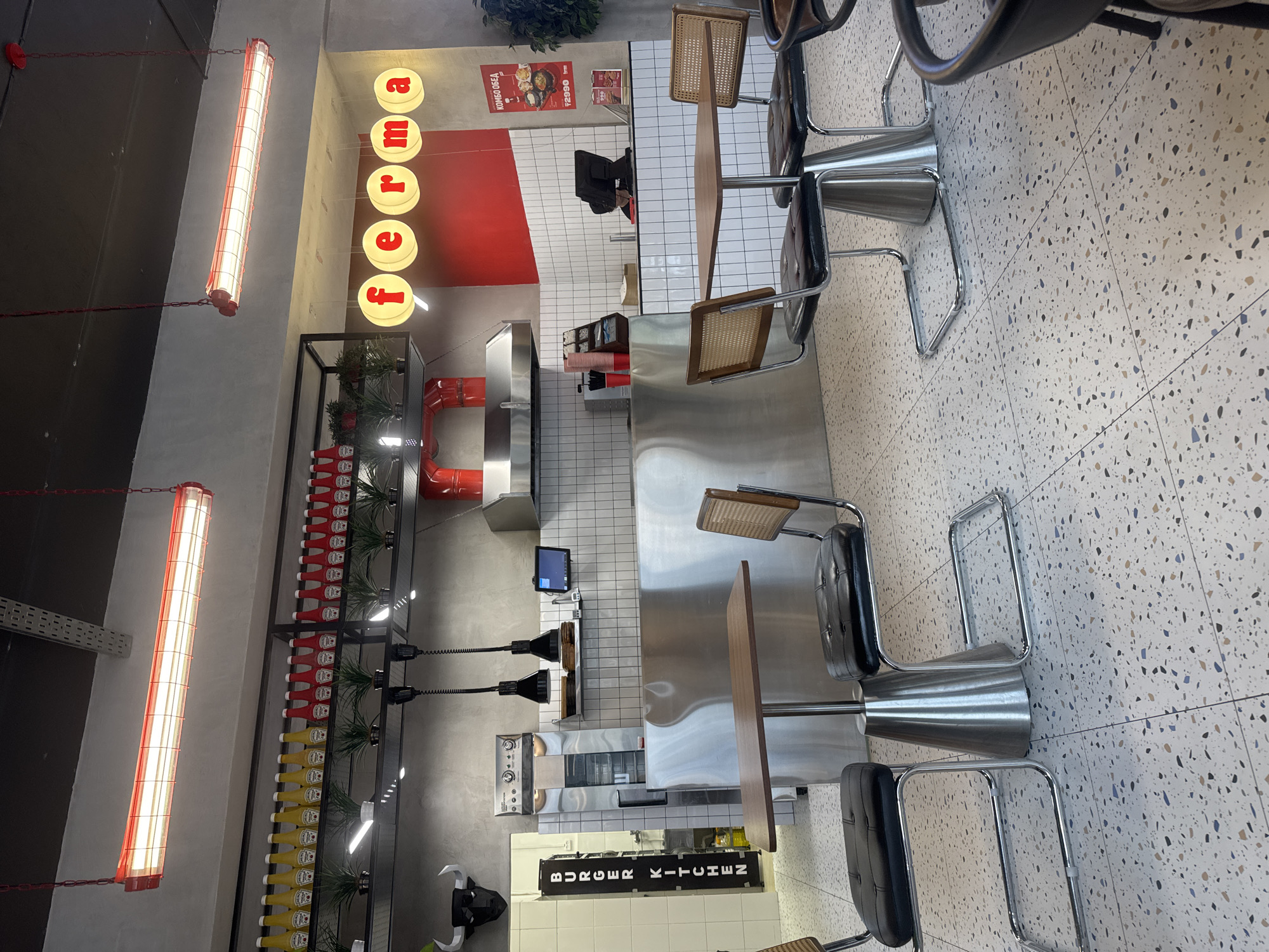 The image depicts the interior of a modern burger restaurant named "ferma" in Kokshetau, Kazakhstan. The establishment features a clean, industrial-chic design with a polished concrete floor, white tiled walls, and stainless steel surfaces. In the foreground, a small wooden table is set with two chairs, each with black leather seats and rattan backs, resting on a chrome base. To the left, a vertical sign reads "BURGER KITCHEN" in white lettering against a black background.
The background reveals a well-equipped kitchen area. A stainless steel counter with overhead shelving displays various condiments, including rows of yellow mustard and red ketchup bottles, interspersed with small potted plants. Above the counter, professional kitchen equipment such as ovens and a range hood are visible. Hanging light fixtures, some industrial black pendant lights and some circular illuminated signs spelling out "ferma," illuminate the space.
On the right side of the image, a menu board displays prices and food items, with a prominent advertisement for a "KOMBO OBED" (combo meal) costing 2990 Tenge. The overall atmosphere suggests a busy, functional, and stylish eatery. No people are clearly visible in the image, though a person may be obscured behind the counter. The scene is brightly lit, suggesting daytime operation.
