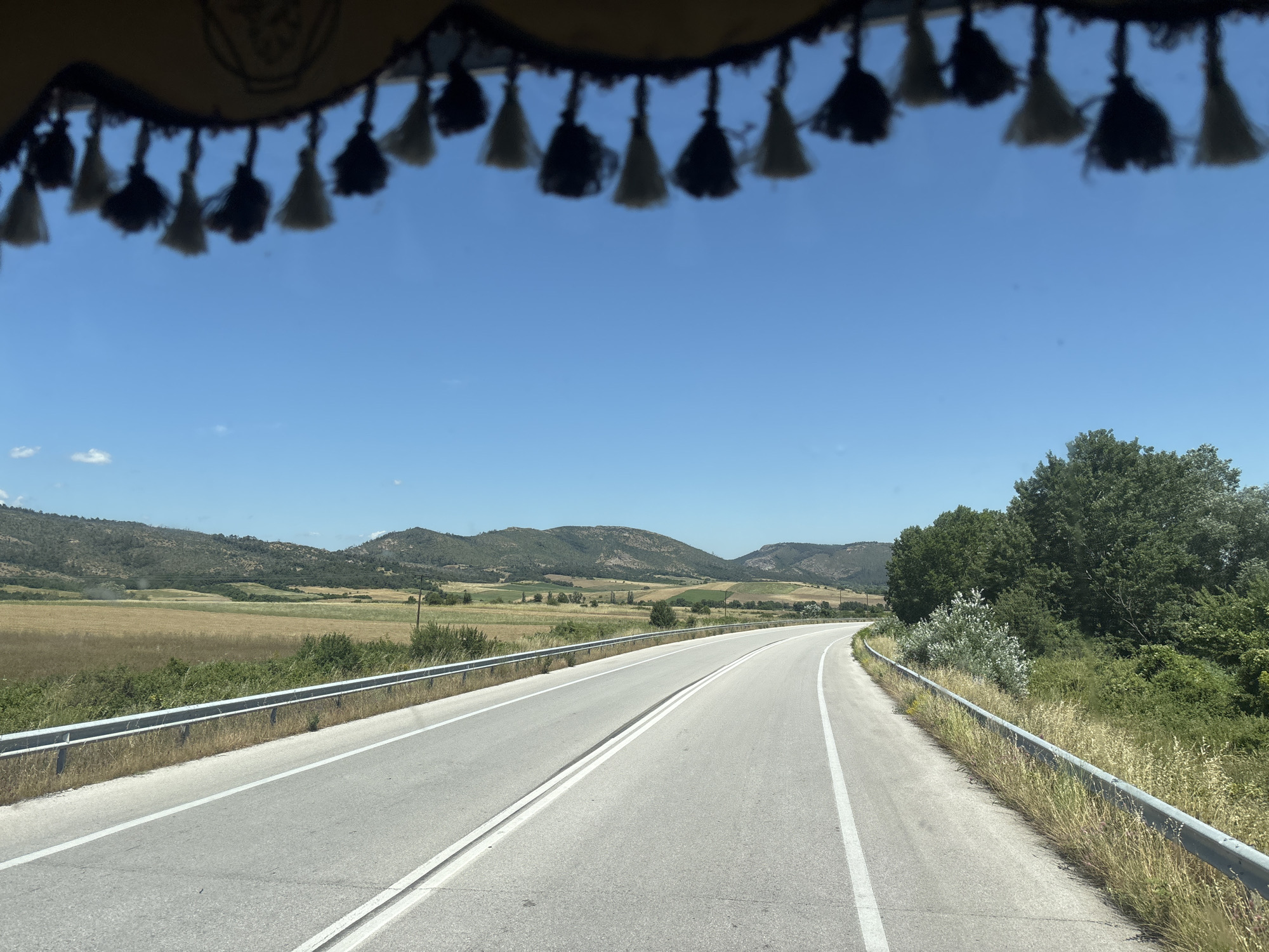 The image shows a view from inside a vehicle, looking out through a windshield that is partially obscured by decorative tassels hanging from the top. The tassels are black and off-white, and appear to be made of yarn or thread.
The scene outside is a rural landscape under a clear blue sky. A paved road curves to the right, with a metal guardrail running along its edge. The road is divided by double white lines. To the left of the road, there is a field of dry grass or crops, and beyond that, rolling hills covered in sparse vegetation and trees. To the right of the road, there are dense green trees and bushes.
The location is identified as Lyra, Greece. The time of day appears to be daytime, likely late morning or early afternoon, given the bright sunlight and clear sky. There are no people or specific actions visible in the image. The environment is natural and appears to be dry and somewhat arid, consistent with a Mediterranean climate.