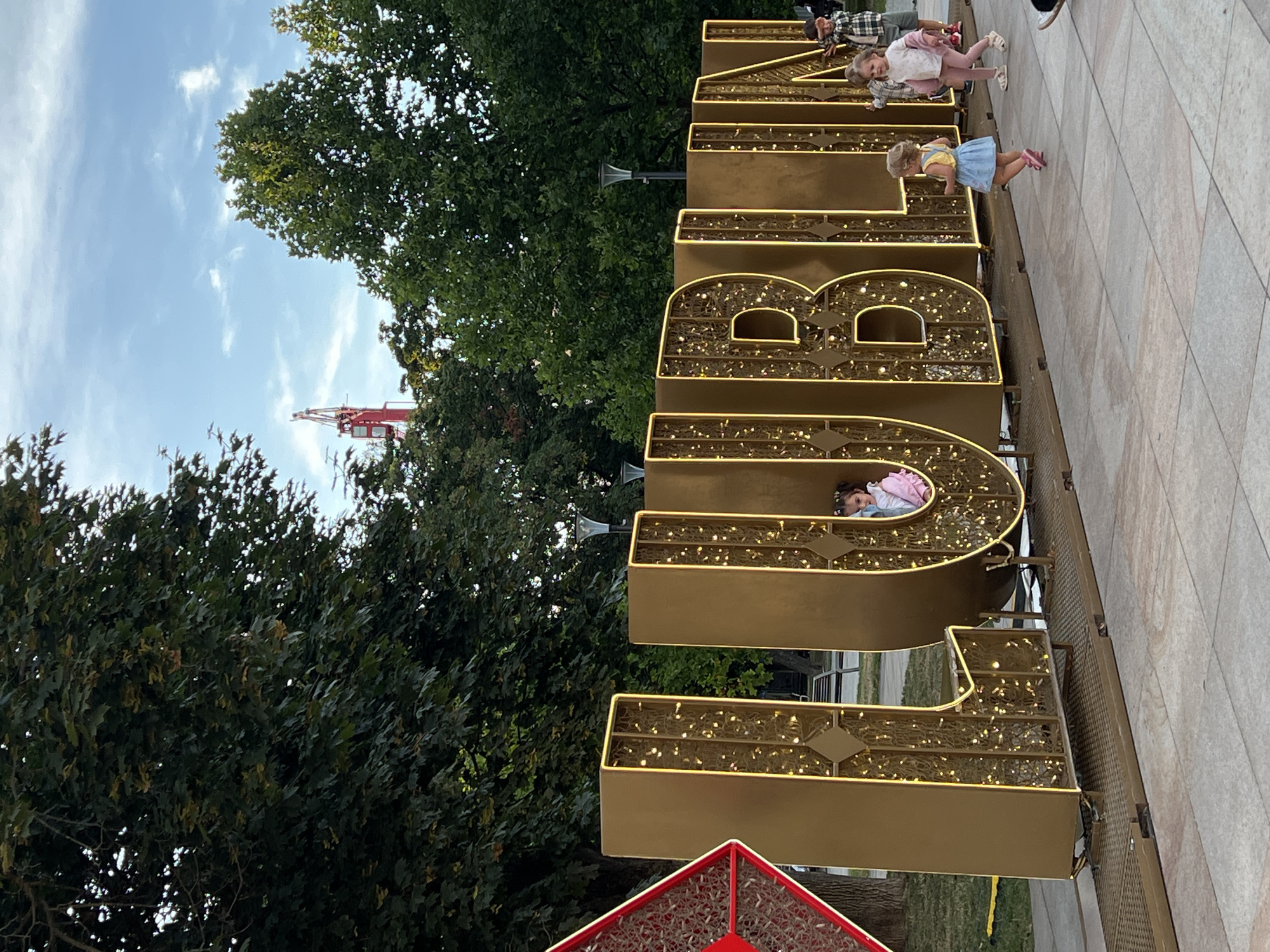 The image depicts a large, golden, illuminated sign spelling out "LUBLIN," the name of the city in Poland. The sign is decorated with numerous small, warm-toned lights, creating a festive and eye-catching display. Several children are interacting with the sign. In the letter "U," a young girl with dark hair is peeking out. To the right of the sign, a young girl in a blue dress is running, and a slightly older girl in pink pants and a white shirt is walking. In the background, there are tall trees, lampposts, and a bright blue sky with some clouds. A red construction crane is visible in the distance above the trees. The ground is paved with light-colored tiles. The overall scene appears to be an outdoor public space, likely a park or plaza, in Lublin.