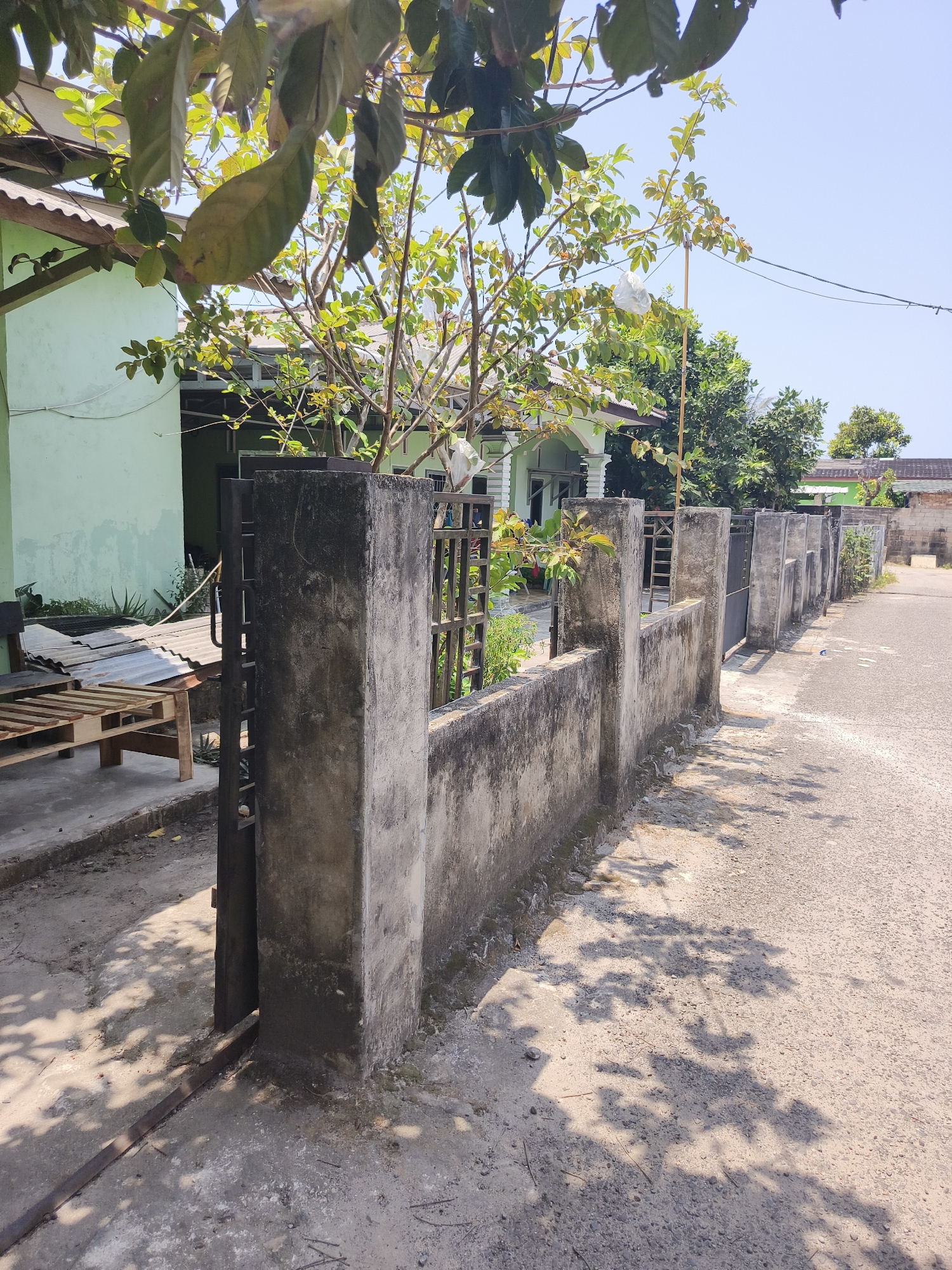 The image depicts a view of a street in the city of Pangkalpinang, Indonesia, on a clear, sunny day. In the foreground, a weathered concrete wall with intermittent fence posts runs along the side of the road. The wall appears aged and shows signs of discoloration and wear. To the left of the wall, a metal gate stands ajar, revealing a glimpse of the property behind it. This property features a light green house with a white-trimmed roof, partially obscured by the branches of a tree with green leaves. In front of the house, a makeshift bench constructed from wooden pallets and corrugated metal is visible. The ground is a mixture of concrete and dirt, with some scattered leaves and debris. The road itself is paved with asphalt and shows distinct shadows cast by the tree and foliage. Further down the street, more houses and vegetation can be seen, along with power lines overhead. The overall scene suggests a residential area in a tropical climate.
