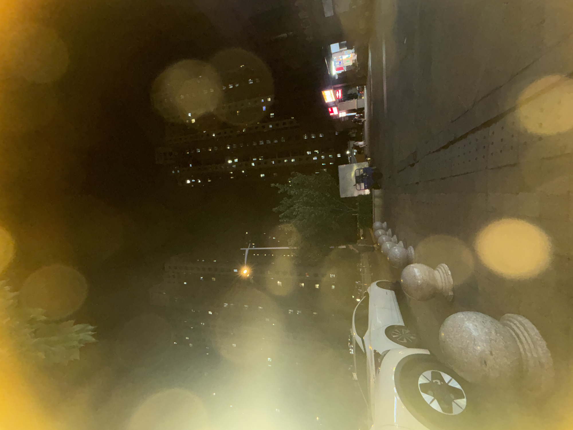The image captures a nighttime scene in a residential area of Suzhou Industrial Park, China. A white car is parked on the side of a paved road, with several stone bollards lining the pavement in front of it. Tall apartment buildings with illuminated windows dominate the background, suggesting a densely populated urban environment. A street light is visible, casting a warm glow. Further down the road, there are what appear to be small shops or stalls with illuminated signs. A few people are walking in the distance. The overall atmosphere is dark, illuminated by artificial lights, with some bokeh effects from the camera's lens.
