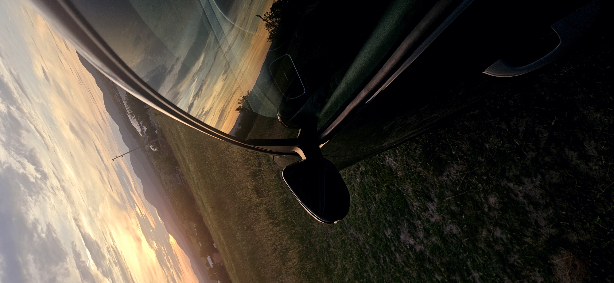 A vertical, low-angle shot captures a car's exterior against a sunset over a rural landscape. The car, predominantly black, is seen from its side, with the passenger door and side mirror visible. The side mirror is angled forward, reflecting the sky and the surrounding scenery. The car's window also mirrors the sunset, displaying warm hues of orange and yellow mixed with streaks of gray clouds.
Outside the car, a grassy field with patches of dirt stretches towards rolling hills in the distance. The hills are dotted with trees and small buildings, suggesting a village or town. A solitary utility pole stands against the sky on one of the hills. The sky itself is a dramatic canvas of a setting sun, casting a warm glow across the clouds and the landscape. The light is fading, creating a contrast between the illuminated sky and the shadowed foreground dominated by the car. The image appears to be taken in the vicinity of Momin Prohod, Bulgaria, given the landscape's characteristics. There are no people visible in the image.