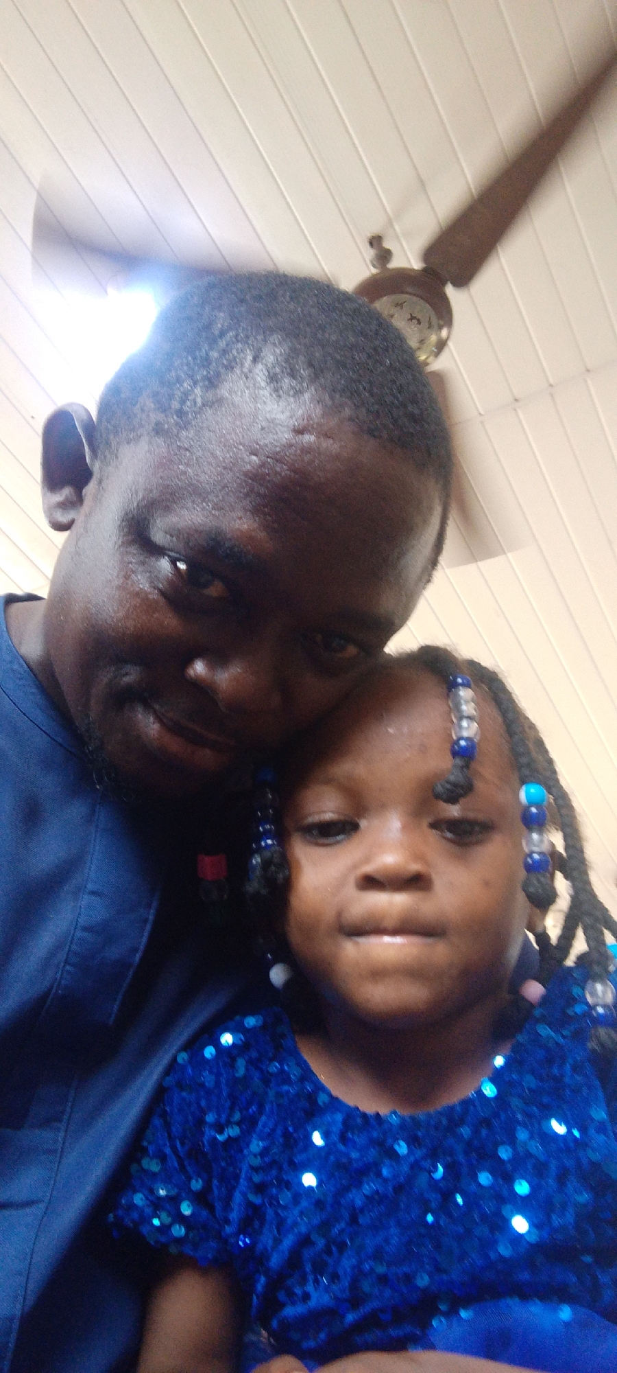 A man and a young girl are posed closely together, likely for a photo, indoors. The man has dark skin, short black hair, and is wearing a blue shirt. The girl, who is positioned slightly in front of the man, also has dark skin and is wearing a blue dress with sequins. Her hair is styled in braids with colorful beads. Overhead, there is a ceiling fan.
The location of the image in Lokoja, Nigeria is not determinable from the image provided.
