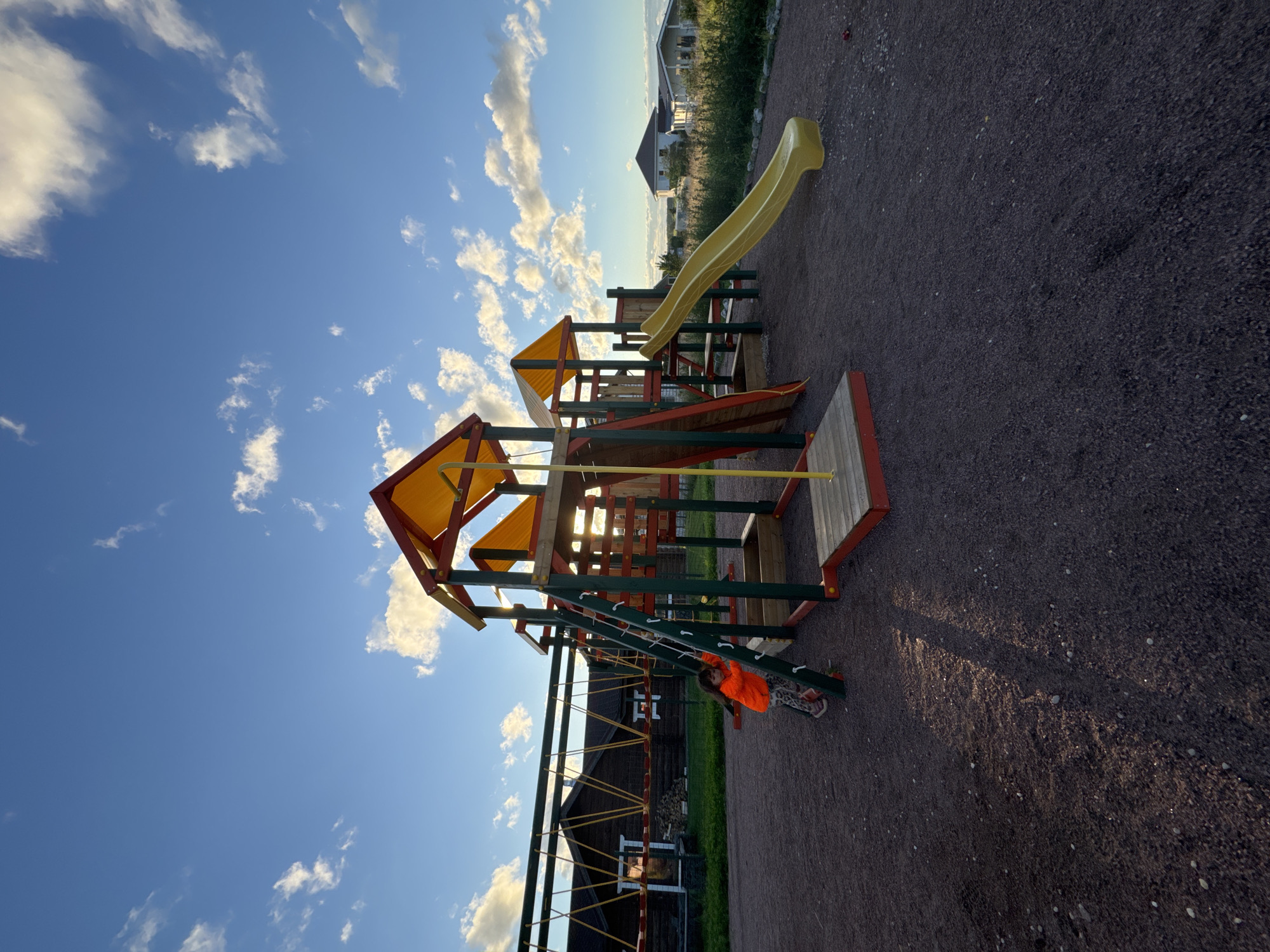 In Kempelevo, Russia, a children's playground is depicted under a bright blue sky with scattered white clouds. The playground features a multi-level structure with a yellow slide, a climbing ladder, and a rope bridge. A young girl in an orange jacket and patterned leggings is seen climbing the ladder on the left side of the structure. The ground is covered in dark gravel, and in the background, several houses with light-colored siding are visible, surrounded by green vegetation. Sunlight casts long shadows across the gravel.