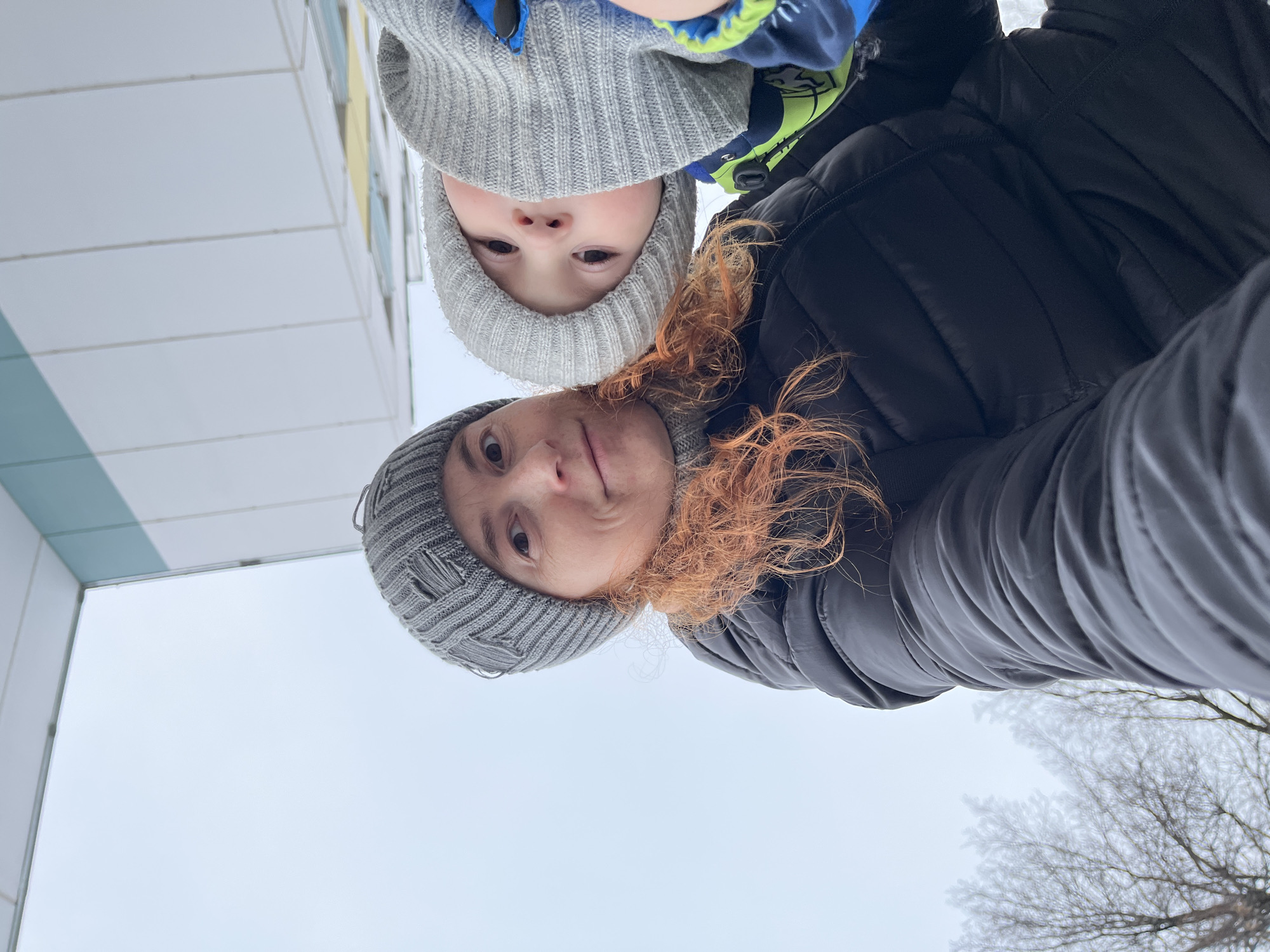 This image presents a unique, low-angle perspective of a woman and a young child, likely a selfie taken from below looking up. Both subjects are positioned at the top of the frame, appearing upside down relative to a standard upright orientation.
The woman, located at the bottom of the visible pair, has striking red or auburn hair, much of which frames her face. She wears a grey knitted beanie and a black puffer jacket. Her expression is a subtle, perhaps curious or slightly amused smirk, with her eyes open wide, looking directly at the camera. Above her, a young child, possibly a toddler, also wears a grey knitted beanie and a blue and neon green winter jacket. The child's face is mostly visible, eyes wide and looking straight ahead with a neutral, curious expression.
The background is dominated by a bright, overcast sky, indicating daylight with diffused light. To the left, the clean lines of a modern building are visible, featuring light-colored panels with hints of yellow and pale blue accents. To the right, the intricate, bare branches of a tree are seen against the sky, suggesting late autumn or winter conditions. The soft focus and slightly hazy appearance around the branches could indicate fog or mist, consistent with cold weather.
The subjects' warm clothing—beanies and winter jackets—reinforces the cold weather setting, likely a winter or early spring day in Chaikovsky, Russia. The image captures a candid, playful interaction between a parent and child, possibly during an outdoor excursion. There is no visible text in the image.