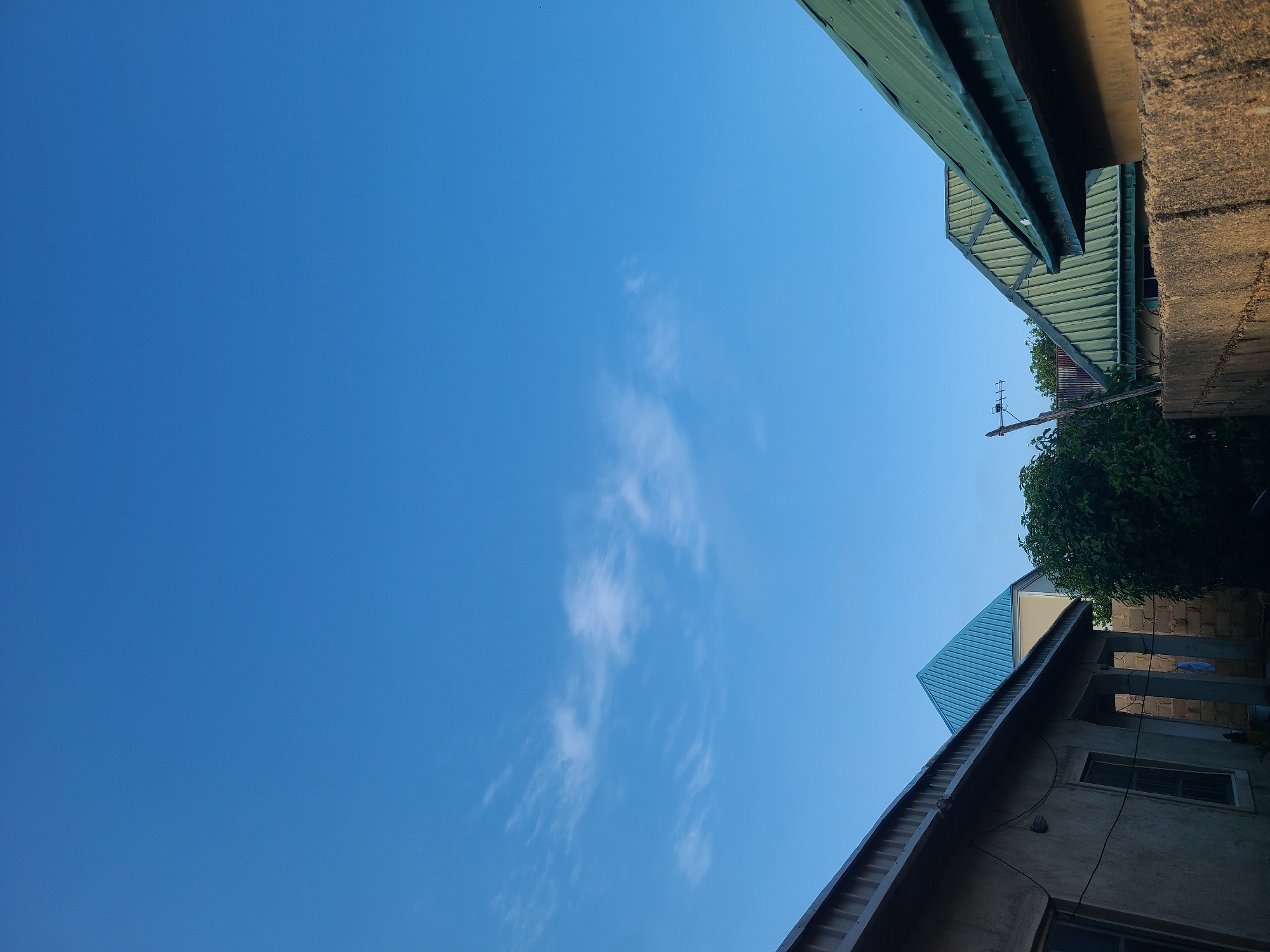 The image captures a clear blue sky above a cluster of buildings with corrugated metal roofs. In the foreground, the roof of a structure extends from the left, made of a lighter colored material, possibly aluminum. To the right, a rough-textured, tan-colored wall runs across the bottom of the frame, with a section of a yellow building visible behind it. The middle ground features two distinct buildings. One on the left has a prominent bright blue, pyramid-shaped roof. To its right, a dense green tree stands tall. In the center-right, a house with a green corrugated metal roof is partially visible, its ridged surface clearly defined against the sky. Another structure with a similar green roof is visible further to the right, also with a ridged pattern. A wooden pole with an antenna is positioned near the tree. The scene appears to be an outdoor residential area in Kuru Karama, Nigeria, under a bright, cloudless sky.