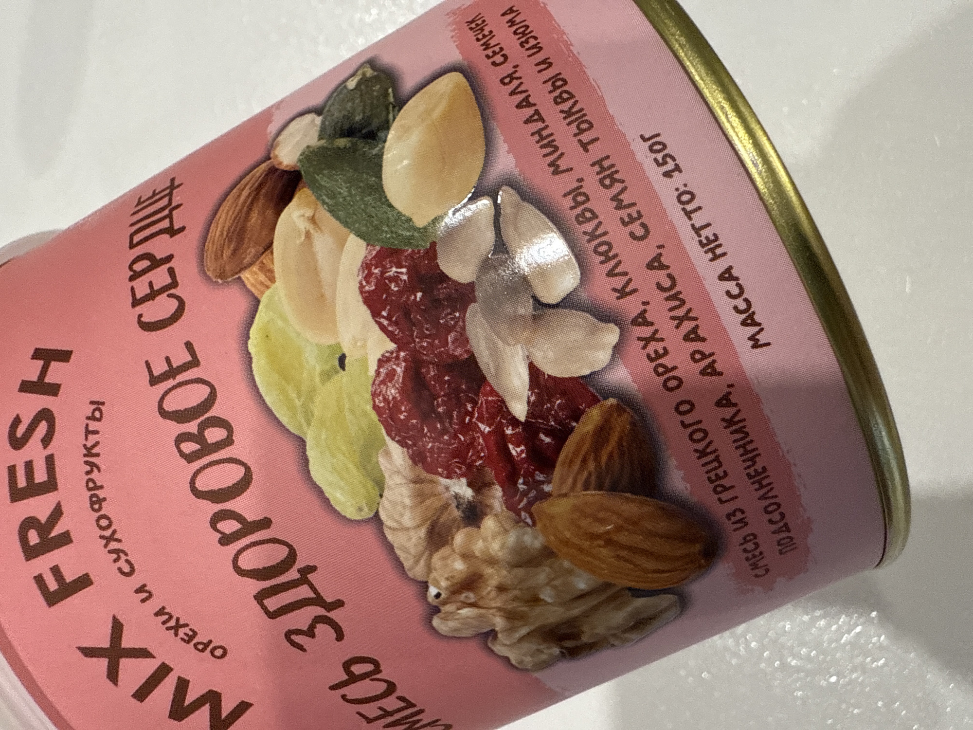 A close-up shot shows a pink can of nut and dried fruit mix. The can is labeled "MIX FRESH" and "HEALTHY HEART" in Russian. The label also lists the ingredients, which include walnuts, cranberries, almonds, sunflower seeds, peanuts, pumpkin seeds, and raisins. The can states the net weight is 150g. There is no indication that this image was taken in Votkinsk, Russia.