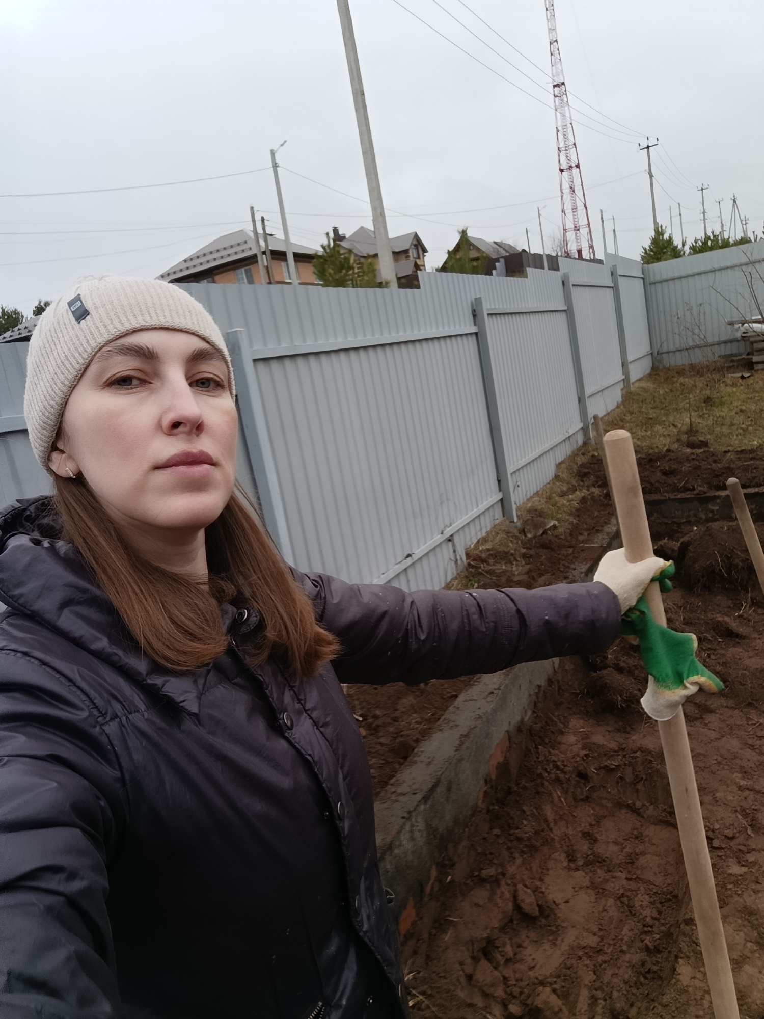 A woman takes a selfie outdoors, looking directly at the camera with a neutral expression. She wears a light-colored (beige or off-white) knitted beanie, a dark puffy winter jacket, and green and white work gloves on her right hand. She is holding the wooden handle of a shovel or a similar digging tool, resting it in the excavated earth beside her.
The scene depicts a plot of land with a freshly dug trench or foundation ditch, revealing brown soil and some concrete or brickwork. A light grey corrugated metal fence forms a boundary in the mid-ground. Beyond the fence, several houses with various roof styles are visible, suggesting a residential area in Chaikovsky, Russia.
The background also features multiple utility poles with power lines strung across them. A tall, distinctive red and white striped communication or radio tower stands prominently on the right side of the frame, towering over the surrounding structures. The sky is overcast and grey, indicating a cloudy day, and the woman's warm attire suggests cool or cold weather, likely late autumn, winter, or early spring. No legible text is visible on her beanie's label. The activity appears to be related to construction, landscaping, or heavy gardening work.