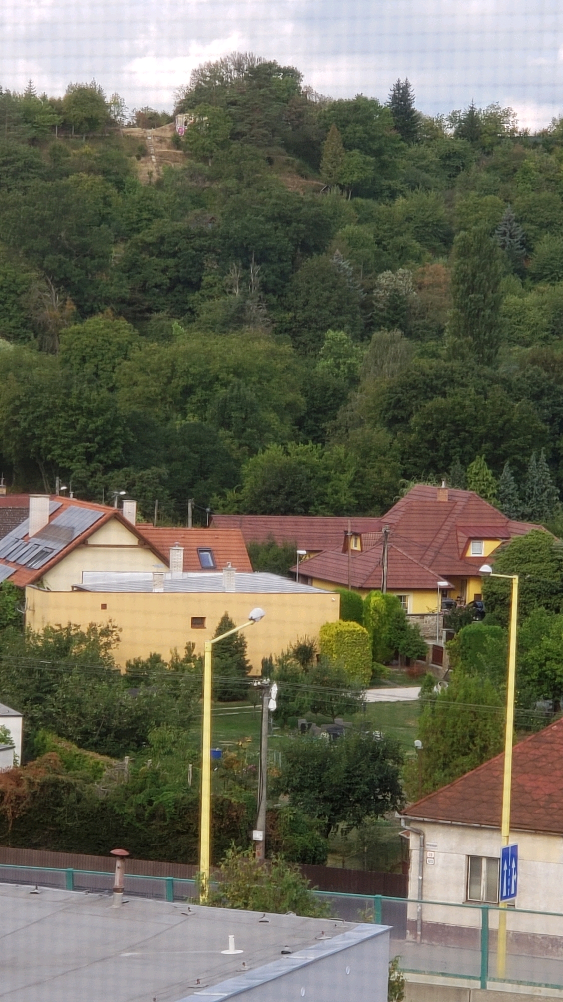 The image depicts a view of a residential area nestled at the base of a wooded hill, likely located in the city of Prešov, Slovakia. The foreground shows the rooftop of a building with a metallic gray surface, featuring ventilation pipes and antenna bases. Beyond this, a cluster of houses is visible. One house is painted yellow with a brown tiled roof, and another adjacent to it has a similar yellow facade and a brown tiled roof. Further to the right, a larger house with a prominent brown tiled roof and white siding is present. These houses are surrounded by lush green trees and vegetation, indicating a suburban or semi-rural environment. A steep, tree-covered hill rises in the background, with a visible path or road leading up to a small structure or monument near the summit, partially obscured by trees and foliage. The sky is overcast with visible clouds. Yellow lampposts are scattered throughout the scene, and a blue and white traffic sign is affixed to one of them. There are no people clearly visible in the frame.