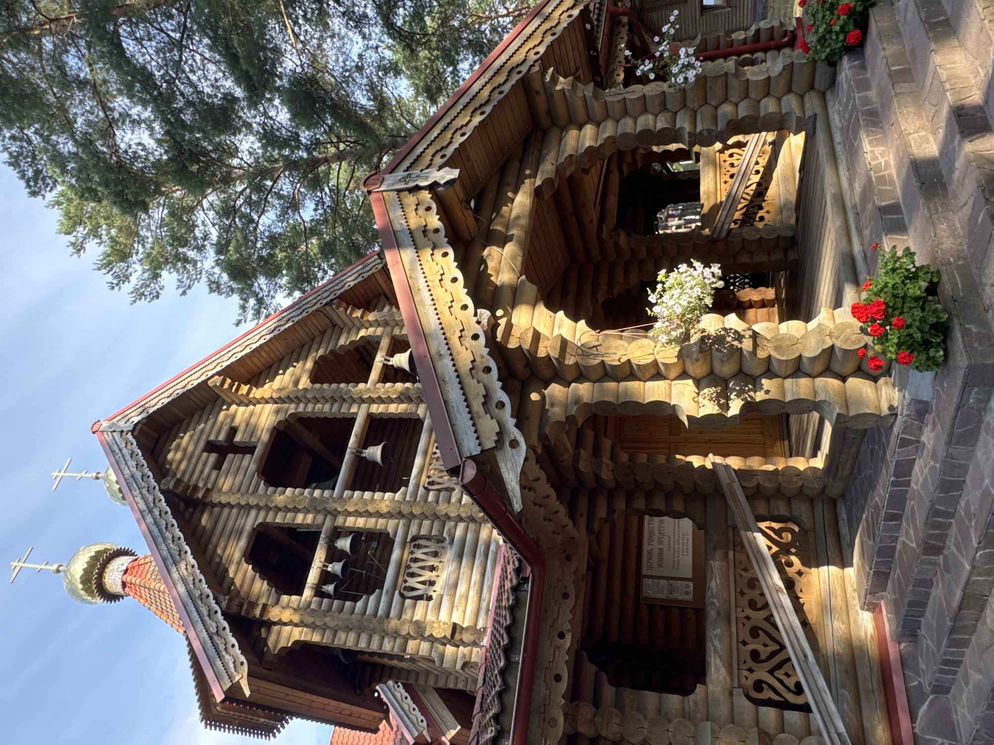 This image shows the wooden Church of the Nativity of the Most Holy Theotokos in Gleybovo, Russia. The church is constructed from logs and features intricate wooden carvings around the rooflines and entrance. It has a traditional Orthodox design with a golden dome topped with a cross. Bells are visible in the upper belfry. In the foreground, stone steps lead up to the entrance, flanked by potted red flowers. A sign with Cyrillic text is mounted on the exterior wall. Tall pine trees are visible in the background against a bright blue sky. The lighting suggests it is daytime, with sunlight casting shadows on the structure. No people are present in the image.