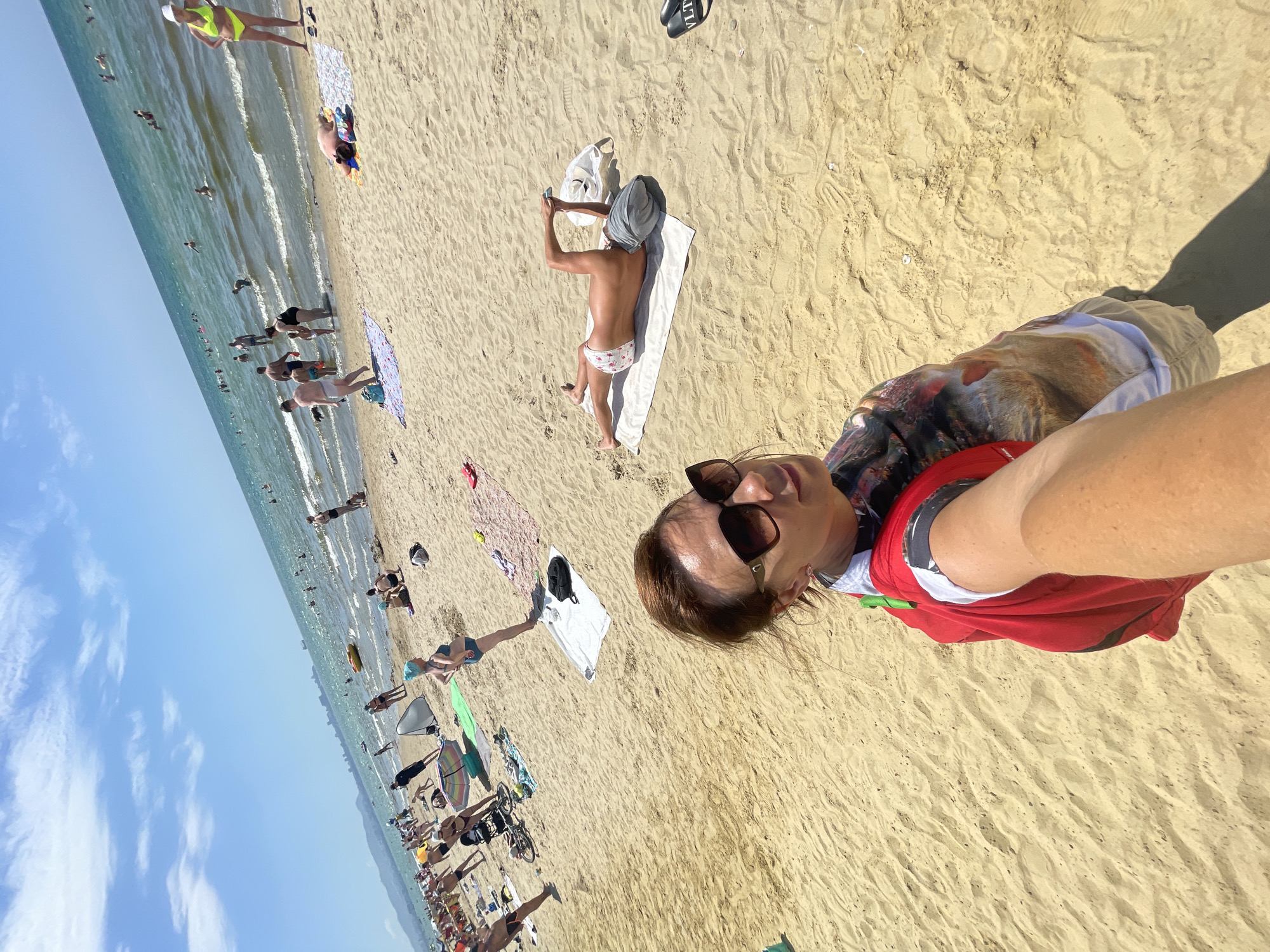 A woman takes a selfie on a sunny beach in Anapa, Russia, with the Black Sea and other beachgoers in the background. The woman is wearing sunglasses and a red garment over a patterned shirt. She is standing on sandy ground, with the sea stretching out behind her. The beach is crowded with people swimming, sunbathing, and relaxing. The sky is blue with a few white clouds. The scene depicts a typical summer day at a popular tourist destination.