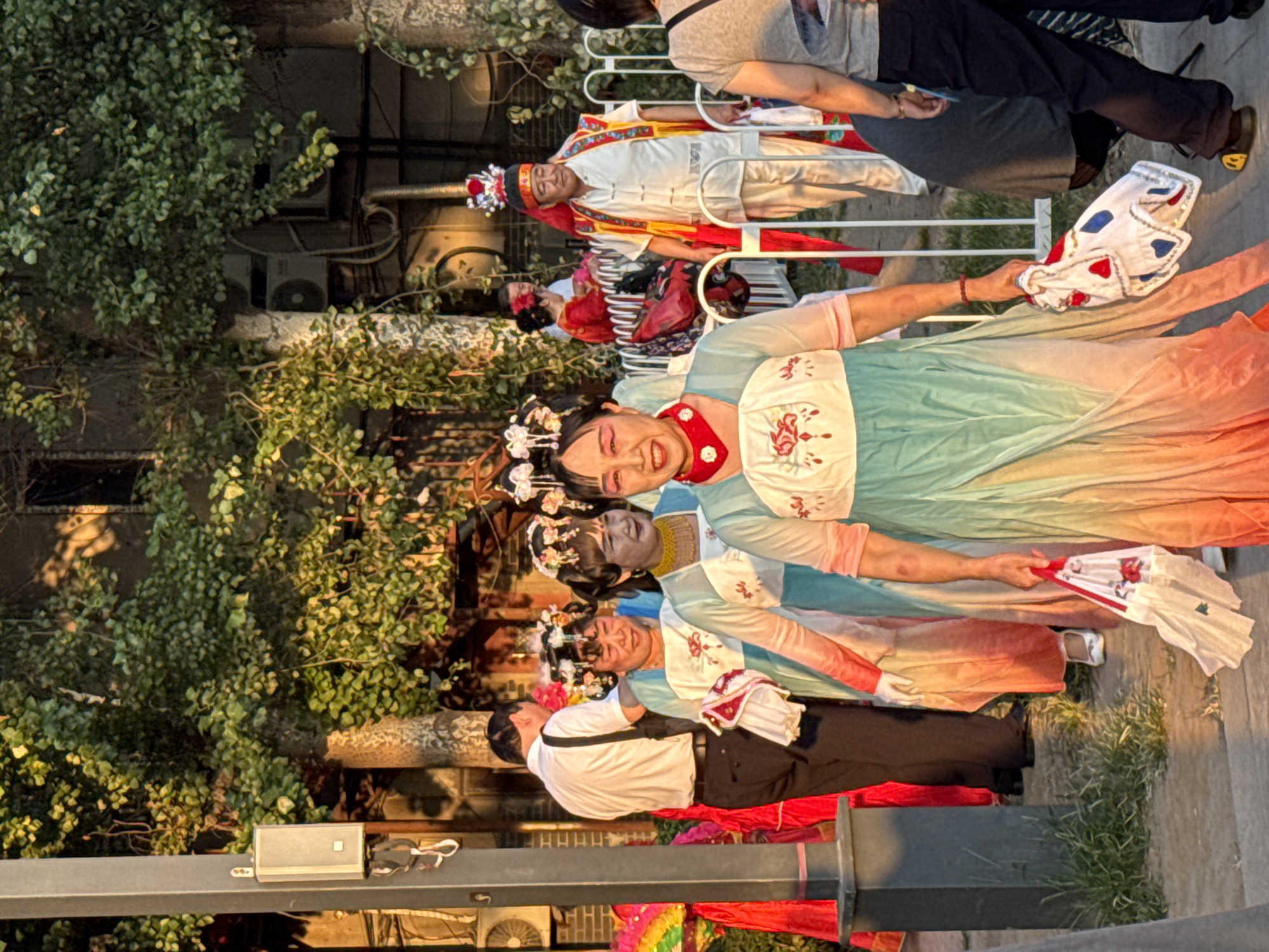 In Jinzhou, China, on a sunny day, a group of people dressed in traditional Chinese opera costumes are walking outdoors. The main subjects are four women in colorful, flowing garments, adorned with elaborate headdresses and face paint. They are smiling and holding white cloths with red and blue accents. To their right, a man in a white outfit and a red cape, also in traditional attire, walks alongside. Behind them, other individuals are visible, including a man in a white shirt and black pants, and people in the background wearing casual clothing. The environment appears to be a public space with trees and foliage, a white metal fence, and a lamppost with a speaker. The lighting suggests it is either early morning or late afternoon, with shadows cast across the scene.