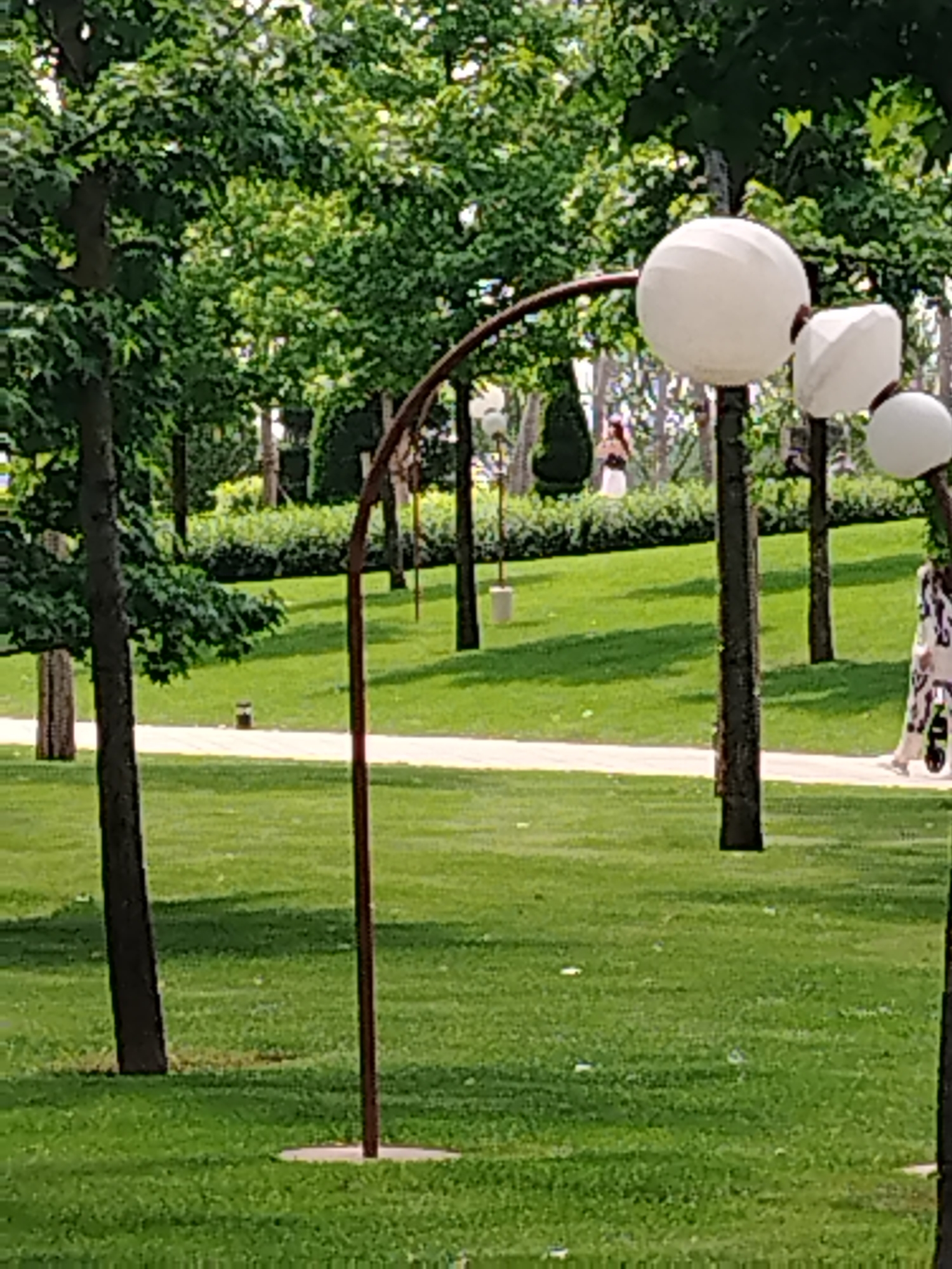 This image depicts a park scene with lush green grass and trees. In the foreground, there is a large, curved metal structure with a circular base. Attached to this structure are several large, white, spherical objects, possibly decorative lights. To the right, a person is walking on a paved path, wearing a patterned dress and pushing a stroller. In the background, another person is visible standing near a sculpted evergreen shrub. The park appears to be well-maintained with manicured lawns and trees providing shade. The overall impression is one of a peaceful outdoor setting. This image was taken in Krasnodar, Russia.
