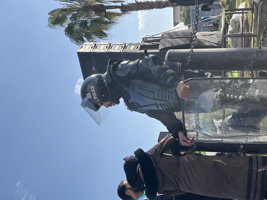 An outdoor scene in Al-Qanawat Municipality, Syria, features a male police officer in riot gear standing in the foreground, facing right in profile. He wears a black helmet with "POLICE" in white, a raised clear visor, and black protective padding on his torso, shoulders, and arms. He has a beard and holds a transparent riot shield with both hands, resting it on a dark metal pole. Behind him to the right, a tall stack of black sound speakers is visible, next to a palm tree. To the left of the officer, the back of a person in a brown fur-lined hooded jacket is partially visible, along with another individual's head. The ground is grassy. In the background, several indistinct people are present on a field, and a light-colored building is partially visible under a clear blue sky with minimal clouds. Metal chains connect dark poles, forming a barrier in the lower mid-ground.