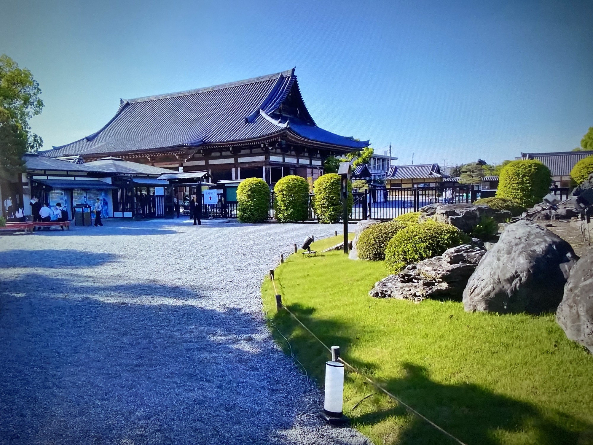The image depicts a traditional Japanese building with a dark tiled roof, situated in an open area covered with gravel. A smaller, adjacent structure is visible to the left of the main building, with people present under its roof. A well-maintained lawn featuring rounded bushes, rocks, and a spotlight, separates the gravel area from the building. A white post connected by a rope serves as a barrier. The location appears to be an Unknown urban setting.