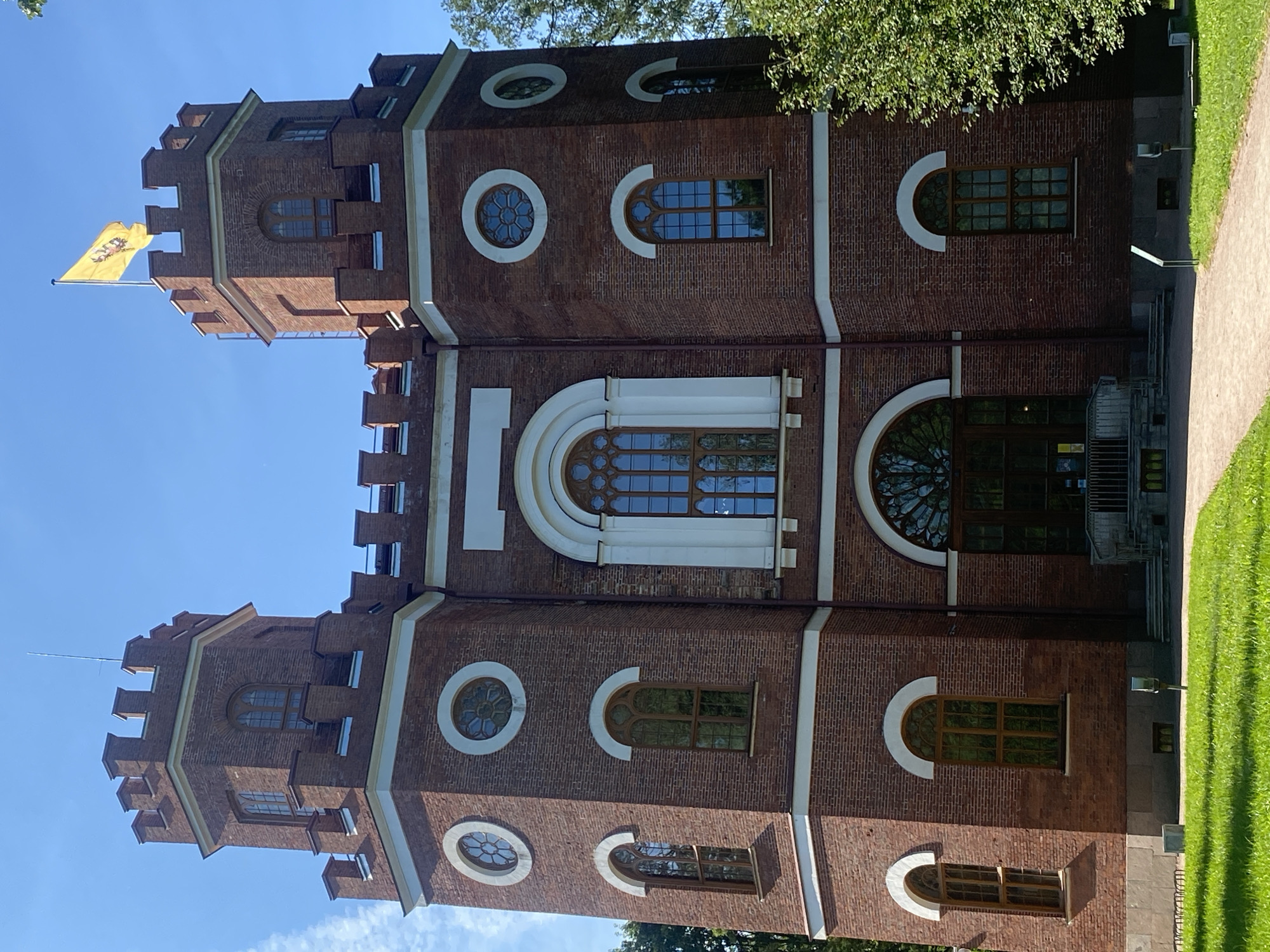 The image shows the Arsenal building in Pavlovsk Park, in the city of Pushkin, Russia. The building is a two-story brick structure with a symmetrical design. It has two prominent towers at the front, each topped with battlements and a flag. The facade is adorned with various windows, including arched windows on the lower level and circular windows on the upper levels. A large, arched entrance with double doors is centrally located on the lower floor. The building is surrounded by lush green grass and trees. The sky is clear and blue. There are no people visible in the image.