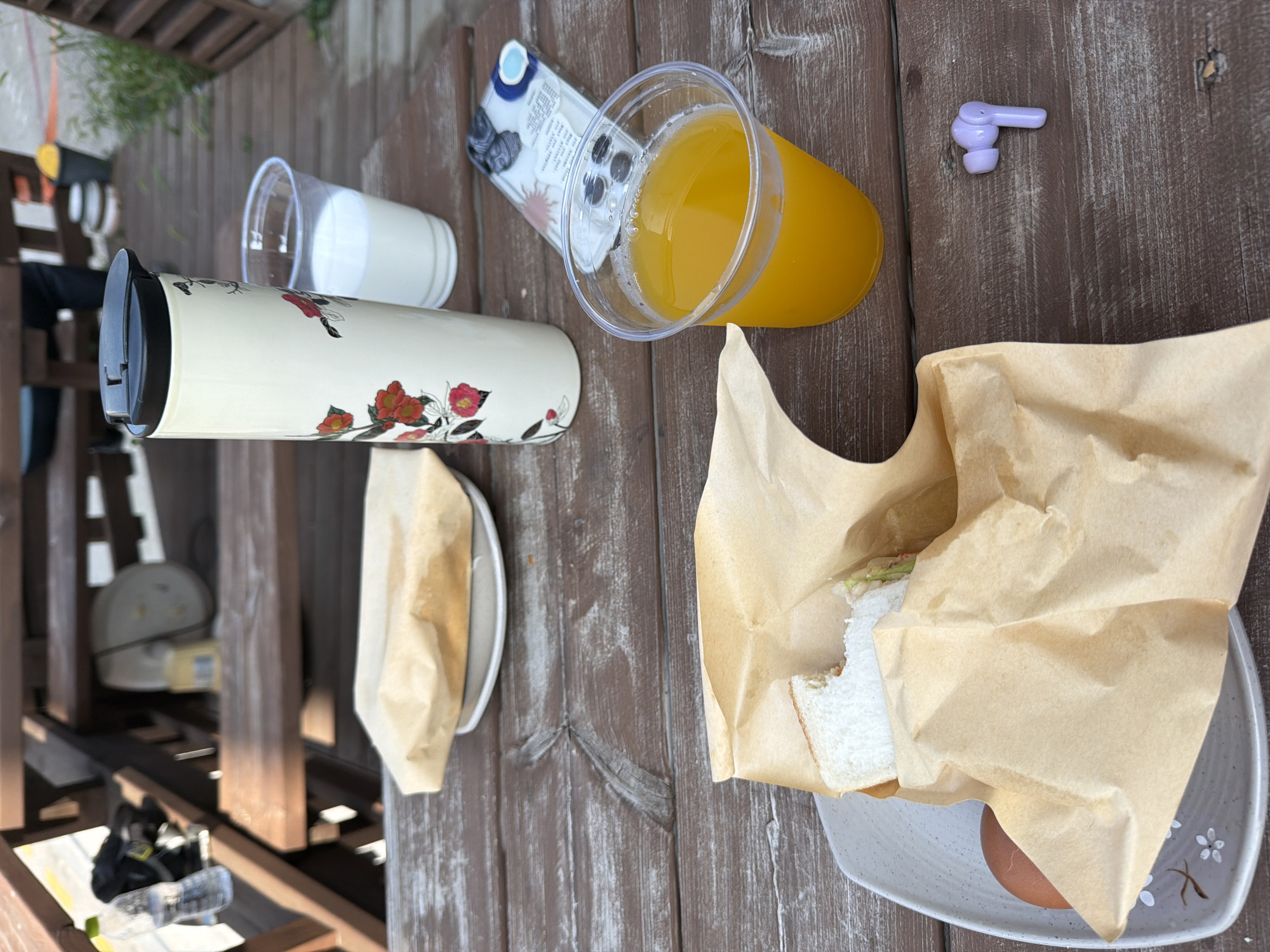 The image captures a casual outdoor meal setting, likely on a wooden table at a cafe or restaurant with outdoor seating. In the foreground, a plate holds an egg and a paper-wrapped sandwich, with a bite taken out of the bread. To the right, a clear plastic cup contains orange juice, and next to it, a single purple wireless earbud rests on the table. In the background, a white travel mug with floral designs stands beside a clear plastic cup filled with milk. Another paper-wrapped sandwich sits on a plate further back. The overall impression is that of a meal being enjoyed outdoors, possibly in Gangneung-si, South Korea, given the context.