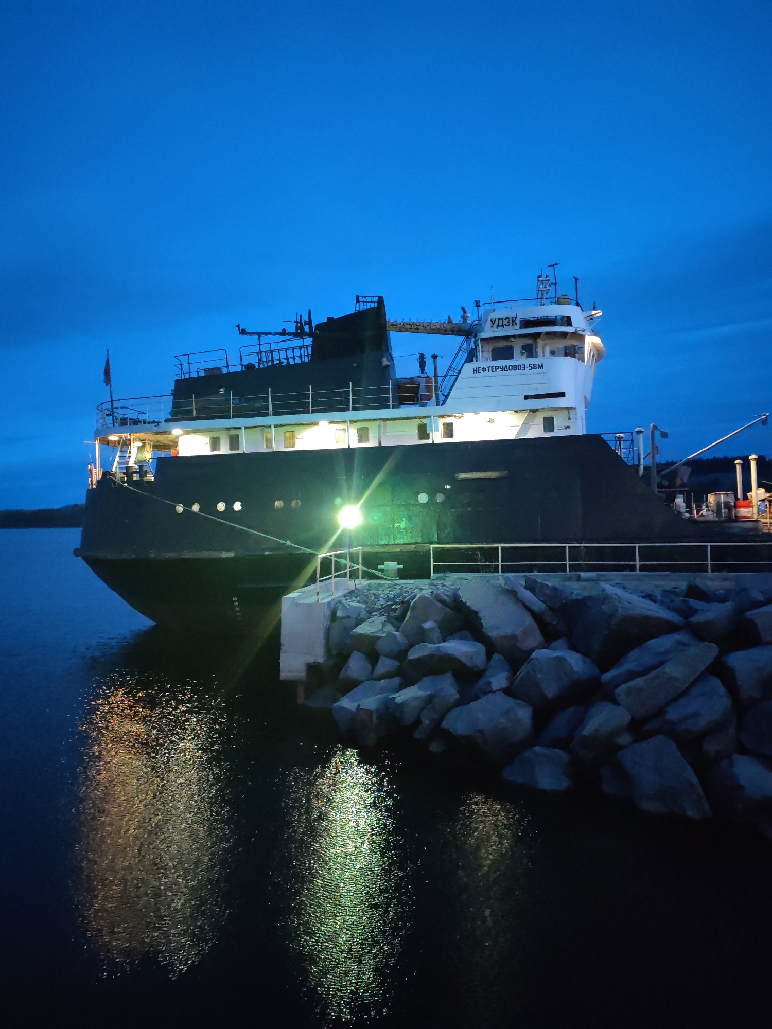 A ship is docked at the port in Shcheleiki, Russia. The ship is large and dark, with a white superstructure on top. The name "NEFTEZAVOD-58M" is printed on the side of the superstructure. The ship is docked next to a concrete pier surrounded by large rocks. The water is dark, reflecting the lights from the ship. The sky is a deep blue, indicating that it is night or dusk. There are no people visible in the image.