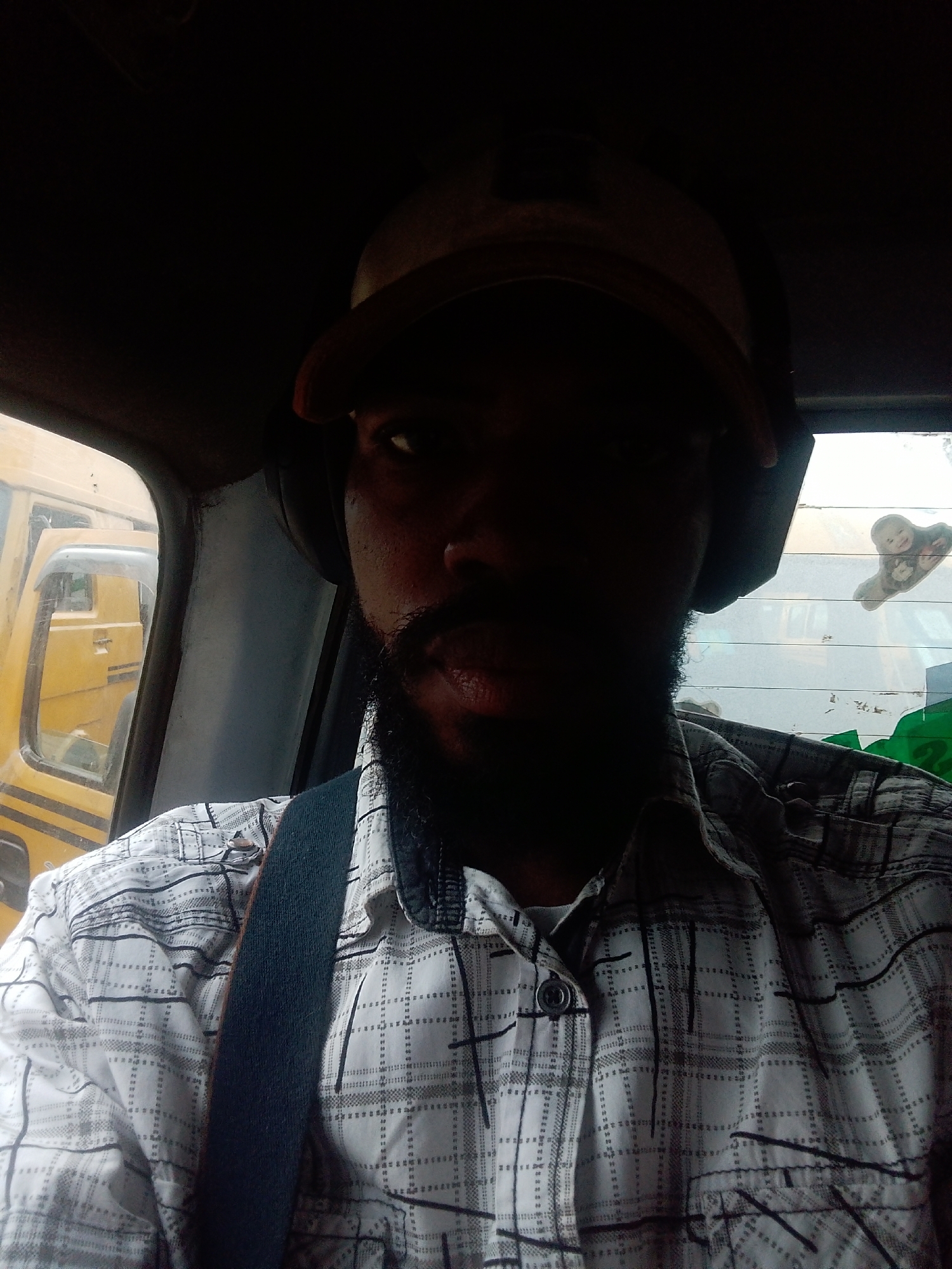The image is a close-up, selfie-style shot of a man sitting in the driver's seat of a vehicle, likely a truck or bus, in Ikeja, Nigeria. The man is wearing a plaid shirt, a baseball cap, and headphones. He has a beard and mustache. The interior of the vehicle is dark, with some light coming from the outside. Through the windshield, another yellow vehicle is visible. There is also a window behind the man with a sticker on it. The scene appears to be a street in Ikeja, given the presence of vehicles and the context.