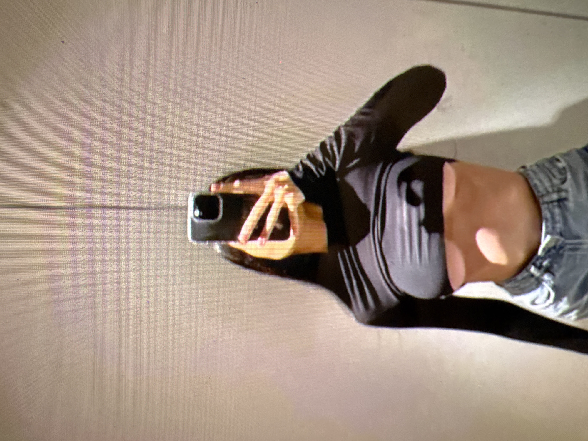 A person is taking a mirror selfie in what appears to be a fitting room with a textured wall. The person is wearing a black long-sleeved crop top that exposes their midriff and a pair of light-wash denim jeans. They are holding up a smartphone to take the picture. The lighting in the room is bright, casting shadows on the person's torso. There are no discernible external features that would indicate the location within the city of Castelo Branco, Portugal.
