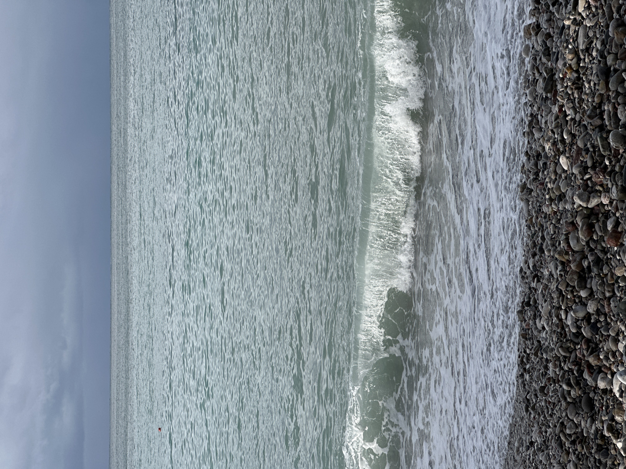 The scene depicts a pebble beach meeting a body of water, presumably the Adriatic Sea, in Šušanj, Montenegro, under an overcast sky. The foreground comprises a dense field of grey, brown, and off-white pebbles varying in size. Moving seaward, a line of white foam marks the immediate waterline, followed by a breaking wave with a prominent white crest and turbulent greenish water. Beyond the breaking wave, the sea surface is a muted teal-green, characterized by numerous small ripples extending towards the horizon. In the upper-mid section of the water, a single small, reddish-orange spherical buoy is visible. The horizon line is flat, separating the sea from a cloudy sky. The sky is dark grey in the upper portion, gradually lightening towards the horizon. No individuals, human actions, or human-made structures other than the buoy are present.