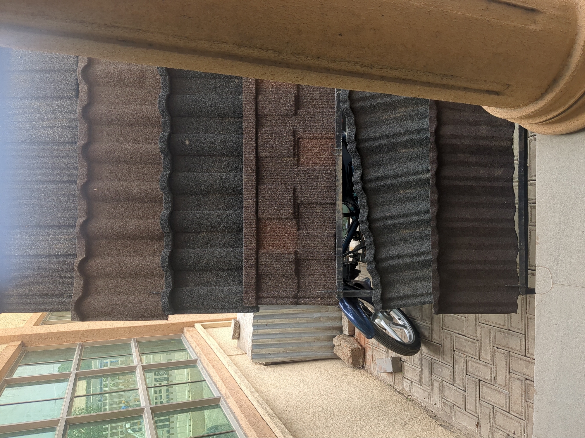 The image shows a collection of stone-coated roofing sheets displayed in a row. They are a mix of dark gray, brown, and a patterned brown variety. A motorcycle, partially visible, is parked behind some of the roofing sheets, with its front wheel and a portion of its blue bodywork in view. The scene appears to be an outdoor display, possibly at a building materials yard or a construction site, given the stacked roofing sheets and the presence of building elements like a window with reflective glass and a textured wall. The lighting suggests it is daytime, but the weather is not discernible. The image is oriented sideways.