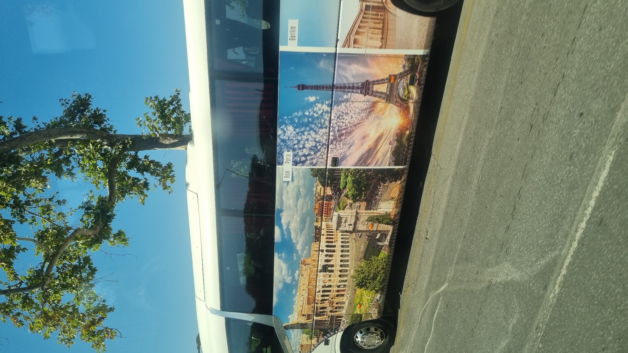 A white tour bus with a wrap showcasing famous landmarks is seen from the side, parked on a paved road with white lane markings. The bus is decorated with images of the Colosseum in Rome, the Eiffel Tower in Paris, and a structure from Berlin. A portion of a tree with green leaves and a clear blue sky are visible above the bus.