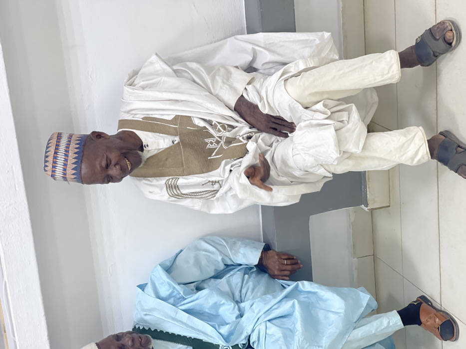 Two elderly men are depicted seated indoors in Zaria, Nigeria. The man on the right, with a dark complexion, wears a white traditional tunic featuring prominent brown embroidery, beige trousers, and a multi-colored geometric patterned cap. He is smiling broadly, exposing his teeth, with his left hand slightly open and extended forward, and his right hand resting on his lap. He wears dark slide sandals. The man on the left, partially visible, also dark-complexioned, wears a light blue traditional garment over a green undergarment and a white cap. A silver ring is visible on the ring finger of his left hand, which rests on his lap. He wears brown slide sandals over black socks. Both men are positioned against a plain, off-white wall with a horizontal relief detail and a grey lower section, above a light-colored tiled floor.