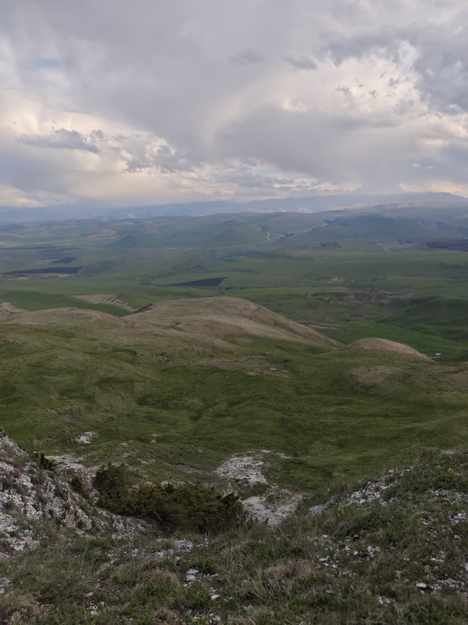 The image captures a landscape in Kichmalka, Russia, featuring undulating green hills in the middle ground and a mountainous terrain in the background. A portion of the foreground is characterized by a rocky, grassy verge with sparse vegetation. The sky is overcast, with cloud formations suggesting a potentially stormy weather. There are no visible people or animals in the image. A faint, narrow road is visible in the distance cutting through the landscape. The lighting conditions suggest it is either early morning or late afternoon.
