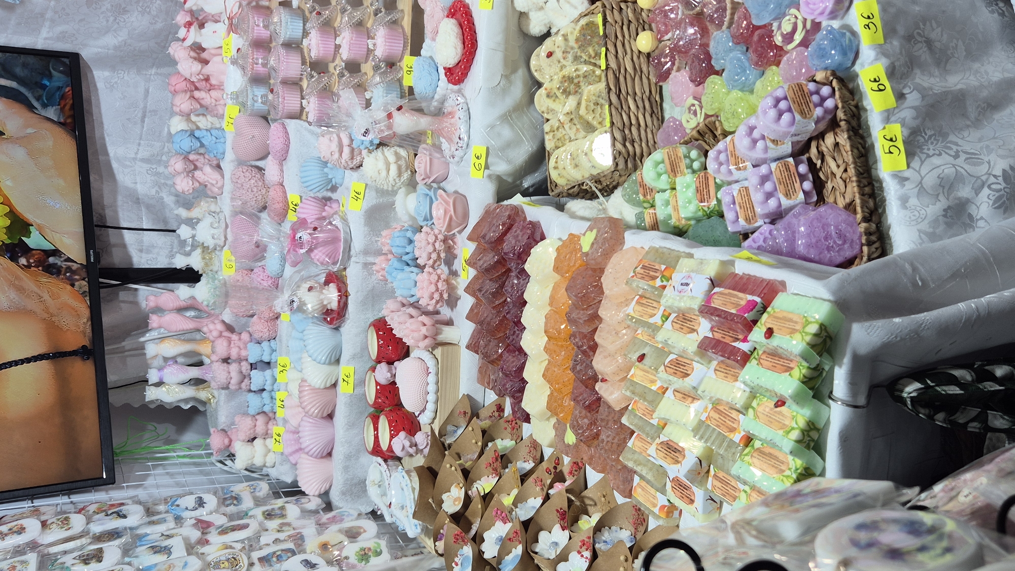 The image shows a stall displaying various artisanal soaps and candles, likely at a market. The items are arranged on white tablecloths on tiered shelves. In the foreground and midground, there are rows of soaps in various shapes and colors, including translucent rectangular soaps in shades of orange, pink, and yellow, as well as opaque soaps in pastel green and cream colors. Some soaps are individually wrapped with decorative labels featuring images of fruits and flowers. In the background, there are shelves filled with decorative candles shaped like teddy bears, dresses, shells, flowers, and cupcakes, predominantly in pink, blue, and white. A television screen is visible at the top, displaying an abstract image. Yellow price tags are placed in front of some of the items, indicating prices such as 7€, 3€, 4€, 5€, and 6€. The overall impression is of a colorful and decorative display of handmade products. The specific location in the city of Primorsko, Bulgaria, is not evident from the image itself, but the presence of price tags in Euros suggests a commercial setting, potentially a tourist market.