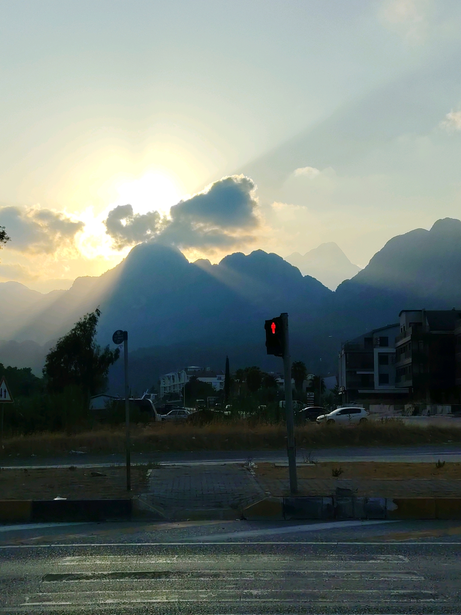 This image, taken in Konyaaltı, Turkey, captures a scene at dusk with mountains in the background. The sun is setting behind a cloud bank, casting dramatic rays of light through the sky and onto the mountains. In the foreground, a pedestrian crossing is visible on a wet road, suggesting recent rain. A traffic light shows a red pedestrian signal. Several buildings, including a multi-story structure, are situated near the base of the mountains. The environment appears to be a developed area with some vegetation, and there are a few vehicles present, though no people are clearly visible.