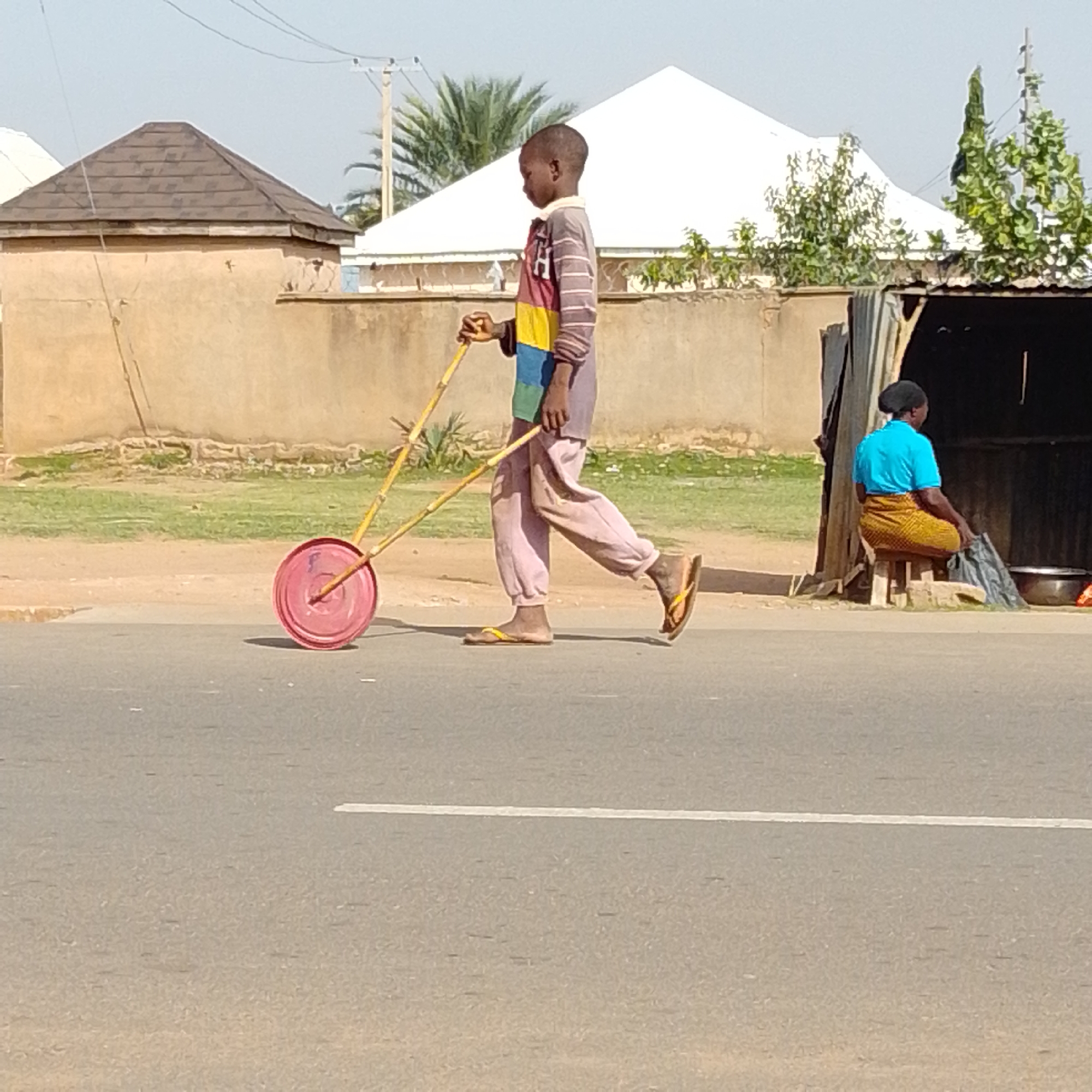 In Tempure, Nigeria, a young boy is captured mid-stride on a paved road, pushing a homemade toy. The toy consists of a red circular object, likely a repurposed lid, mounted on a yellow wooden frame with two handles. The boy wears a striped long-sleeved shirt in muted colors and light pink trousers, along with yellow flip-flops. His posture suggests active engagement with the toy, indicating he is playing.
Behind the boy, the environment transitions from a dusty roadside to a grassy area, bordered by a low, beige-colored wall. Further back, several structures are visible, including a building with a brown tiled, pyramid-shaped roof and a white, steeply pitched roof. To the right, a temporary stall or shelter made of corrugated metal is set up. Underneath this shelter, a woman is seated on a wooden stool, wearing a bright blue top and a patterned skirt. She appears to be observing her surroundings or engaged in an activity, with a black plastic bag and a metal pot nearby. The overall scene depicts a typical day in a Nigerian urban or semi-urban setting, with elements of daily life and children's play.