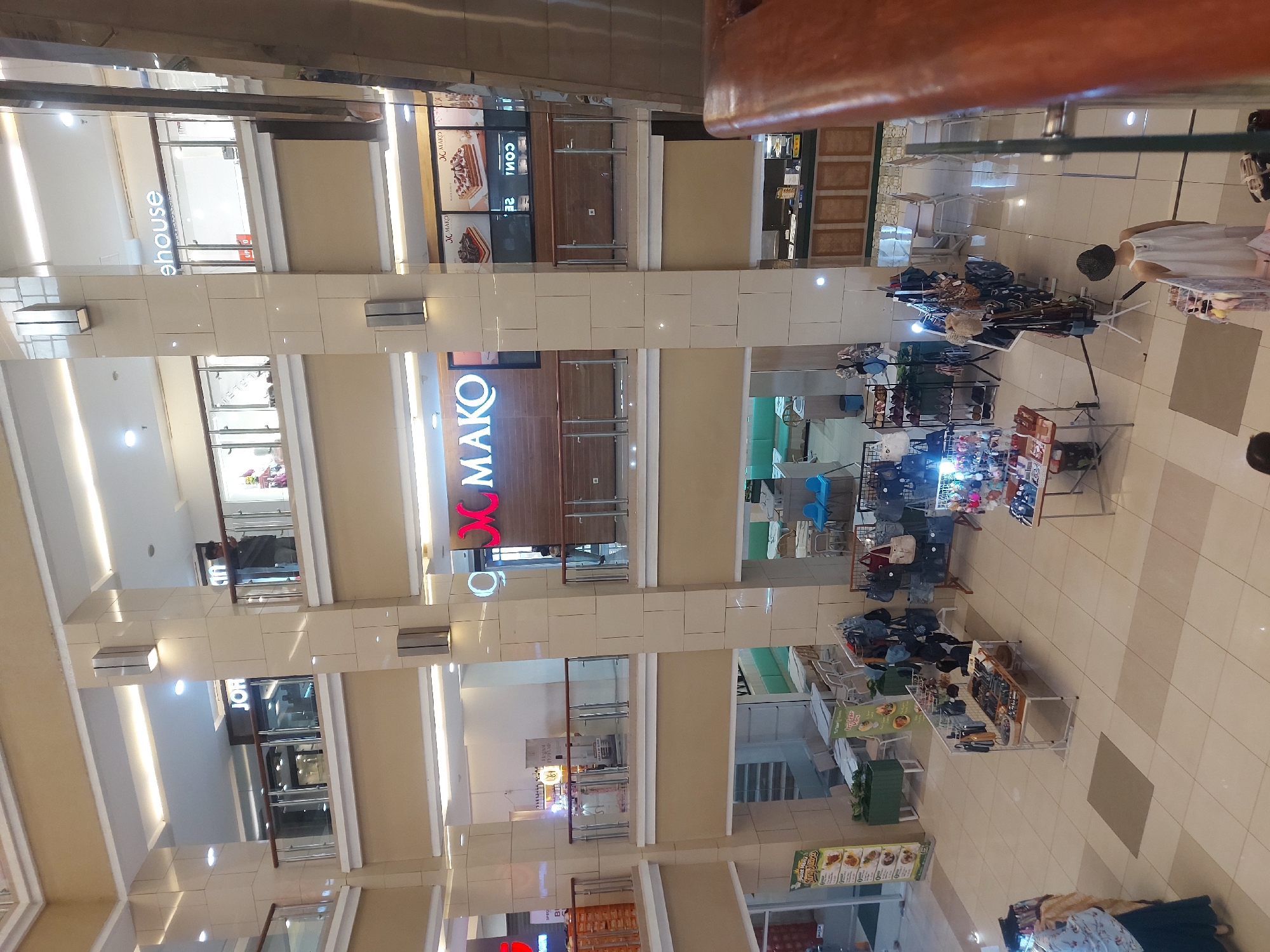 The image shows an interior view of a multi-level shopping mall, possibly in Yogyakarta, Indonesia, with a focus on retail spaces and pedestrian walkways. On the upper levels, storefronts with illuminated signage are visible, including "JOH," "house," and "MAKO." The "MAKO" store features displays of cakes. Glass railings delineate the edges of the upper floors, overlooking the lower levels.
The ground floor or lower level is populated with various retail stalls and a cafe area. Several racks of clothing and bags are displayed, indicating a street market or temporary pop-up shops. One stall exhibits numerous handbags and small accessories. Another stall has clothing items displayed on hangers. In the background of the lower level, a cafe area with tables and chairs is set up.
Several individuals are present in the scene. In the upper levels, a person is seen near the "JOH" storefront, and another is visible in the "house" store. On the lower level, a person wearing a light-colored dress and a hat is browsing at a stall. Another person is partially visible on the far left, appearing to be a shopper. The mall's interior architecture features prominent beige-colored pillars and cream-colored tiled flooring. Natural light seems to be augmented by overhead lighting.