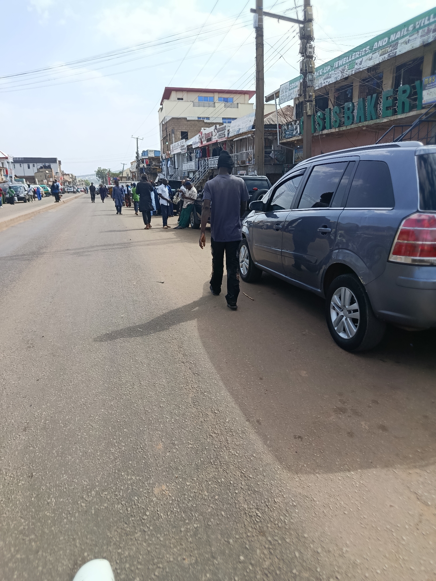 The image depicts a street scene in Jos, Nigeria, with a focus on the road and surrounding buildings. In the foreground, a dark-colored SUV is parked on the right side of the road, partially obscuring the view of the sidewalk and buildings behind it. A man in a dark t-shirt and black pants is walking away from the camera on the right side of the road, casting a long shadow.
The road itself is paved and appears to be a busy thoroughfare with several vehicles and pedestrians visible further down. On the left side of the road, there are buildings that appear to be shops, with awnings and signs. Some of these signs are partially obscured by the vehicles and people.
In the distance, more buildings and people can be seen, suggesting a commercial or residential area. The sky is overcast, and the overall lighting suggests it is daytime. The scene conveys a sense of everyday activity in an urban environment.