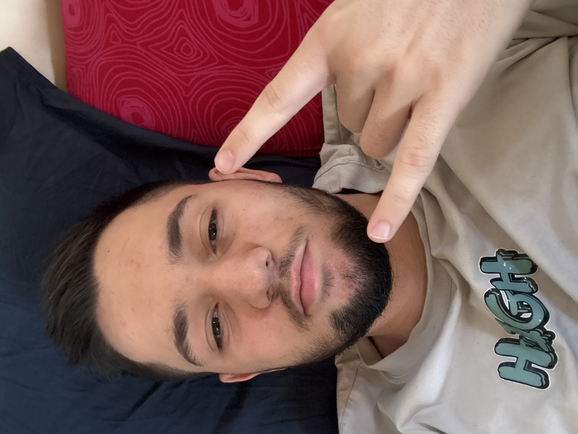 The image shows a young man lying down, captured from an overhead perspective, likely in a bedroom setting in Sofia, Bulgaria. He has dark hair, dark eyes, and a well-kept beard and mustache. The man is looking directly at the camera with a neutral to slightly contemplative expression.
His head rests on a dark blue sheet or blanket, which partially covers what appears to be a red pillow or cushion with a distinct abstract, swirling pattern. The man is wearing a light-colored, possibly beige or light grey, hooded sweatshirt. On the front of the sweatshirt, the word "HIGH" is visible, stylized in a prominent green/blue font with a dark outline.
A hand, presumably the subject's, is making a "rock on" or "devil horns" gesture, with the index and pinky fingers extended, and the thumb holding down the middle and ring fingers. This hand is positioned above his head, with the extended index finger pointing towards his ear. The lighting suggests an indoor environment, but the specific time of day is not discernible.