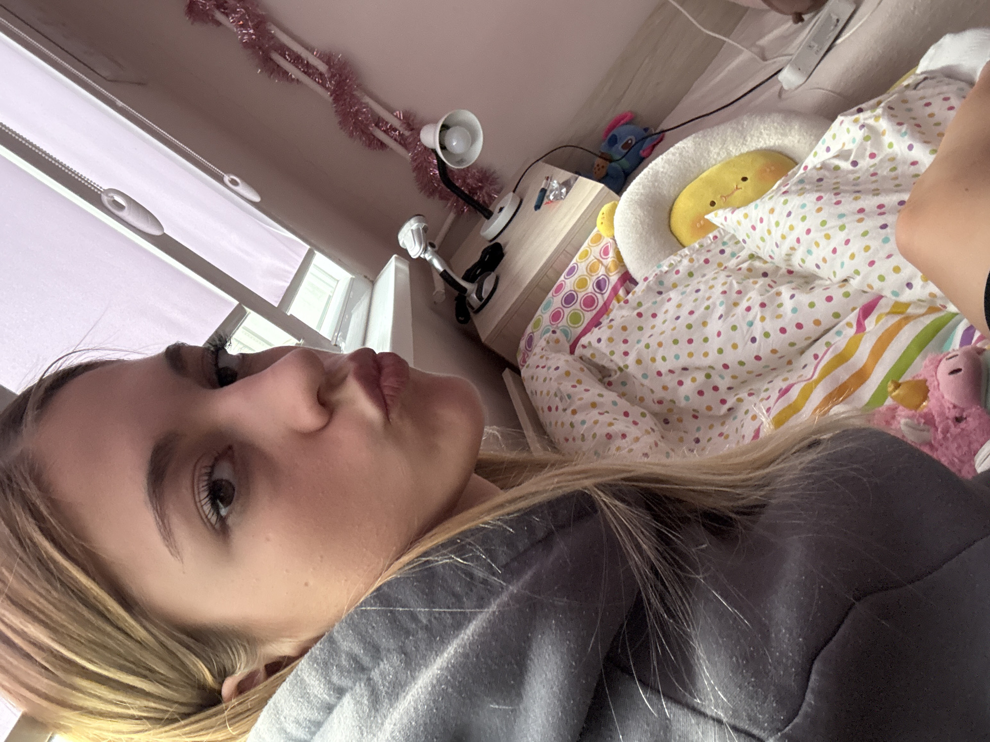 A young person, likely female, is shown in a close-up, upward-angled portrait, possibly a selfie, taken indoors. Her blonde hair is parted on the side, framing her face. She looks towards the camera with dark eyes and her lips are slightly pursed in a neutral to playfully pouting expression. She is wearing a soft, gray garment, likely a sweatshirt or hoodie.
The background reveals a personalized bedroom setting. To the left, white window blinds with pull cords diffuse natural light. Along the wall or furniture above the bed, a pink tinsel garland is draped. A light-colored wooden bedside table or shelf is visible, holding two desk lamps with flexible necks, one white and one black and white. A small blue plush toy, resembling Stitch from Lilo & Stitch, is also on the table.
The bed features colorful bedding, including a polka-dotted duvet or comforter and a striped sheet in vibrant hues. A round, white furry cushion holds a yellow, oval-shaped plush toy with small black eyes and a small mouth. Another small, pink plush toy is partially visible near the foot of the bed. The lighting suggests it is daytime. The overall scene depicts a casual moment in a cozy room located in Sofia, Bulgaria.