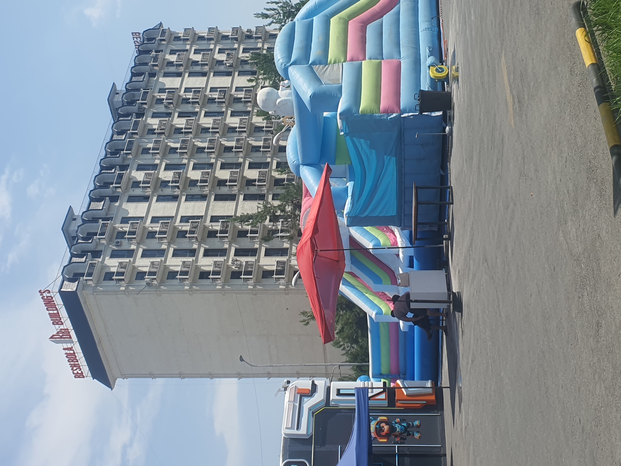 The image shows a bright, sunny day in Fergana City, Uzbekistan, with a clear blue sky and some scattered clouds. The scene is set outdoors, next to a large, multi-story building. To the right of the building, there is a large, inflatable playground structure with bright blue, pink, and green sections. A red umbrella is set up in front of the inflatable, and a few people are visible near a table and chairs, possibly taking a break or supervising. Further to the right, a portion of a street or parking lot is visible, marked with yellow and black bollards and a yellow line. In the foreground, there's a glimpse of a decorated bus or truck with cartoonish figures on its side. The building in the background appears to be a hotel or office building, with many windows and air conditioning units. The text "BESHBOLA BUILDING'S" is visible in red letters on the side of the building. The overall impression is of a lively, possibly recreational, area during the day.