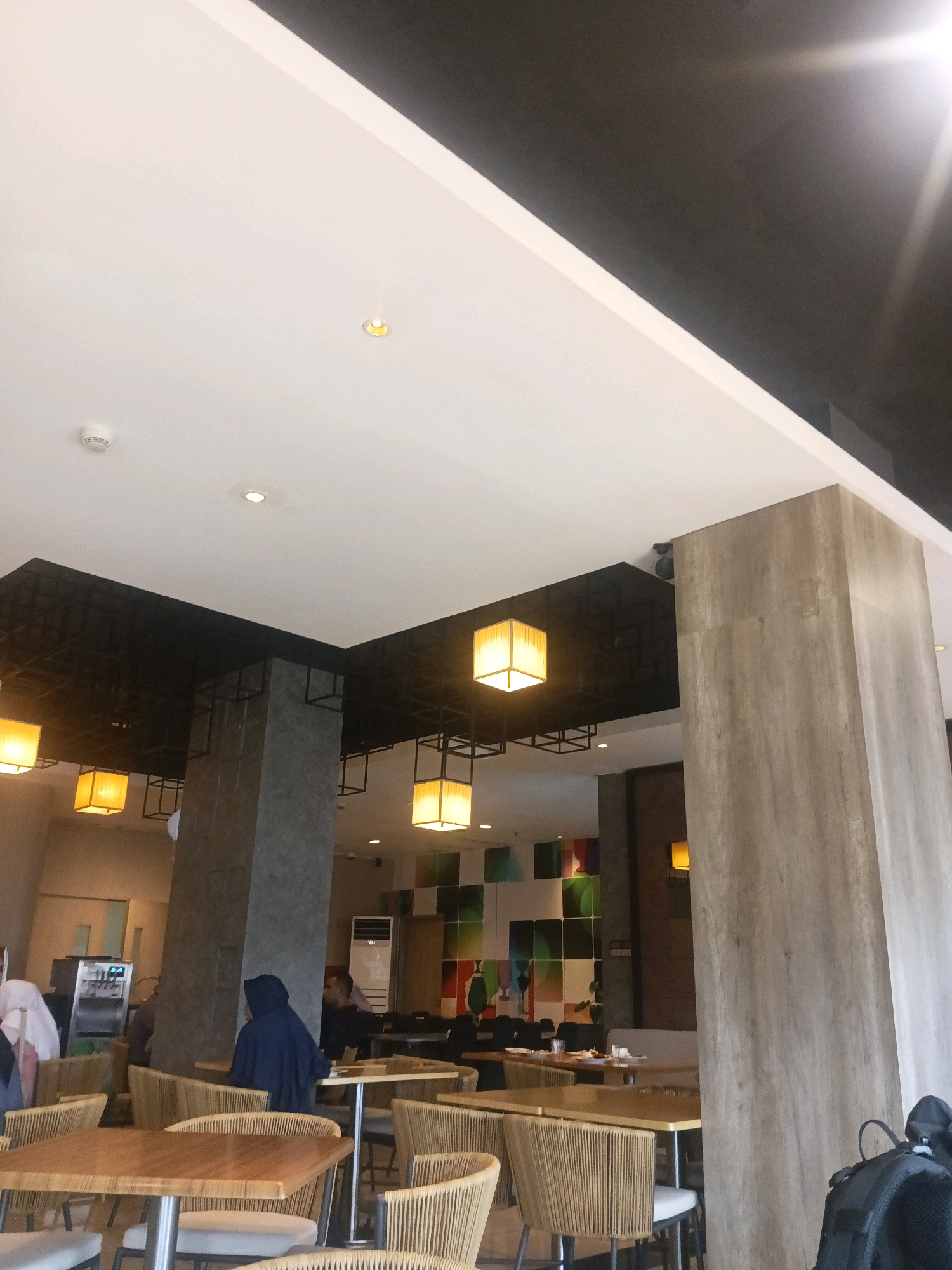 The image depicts the interior of a modern cafe or restaurant. The space features high ceilings with both white and dark grey sections. Several square, yellow pendant lights hang from the ceiling. The interior is furnished with wooden tables and chairs with woven backs, arranged in a casual dining setting. Large, grey, rectangular columns with a wood-like finish are strategically placed throughout the space. In the background, there is a colorful, abstract art installation on the wall. A few individuals are visible, including a person wearing a dark blue hijab and another person in a black shirt. There's also a glimpse of a water dispenser or coffee machine on the left side. The lighting suggests it is daytime, with natural light possibly entering from unseen windows. The overall impression is a contemporary and inviting dining environment. This establishment is likely located in Surabaya, Indonesia, given the typical cafe architecture and decor found in urban areas of the city.