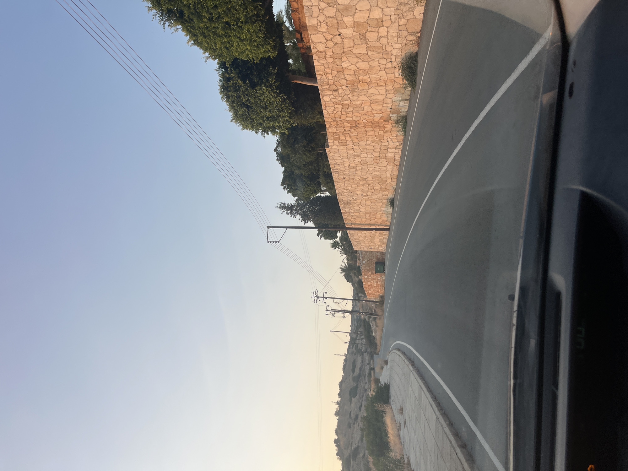 A paved road curves through a semi-arid landscape in Agios Tychona Community, Cyprus. The road is bordered on the left by a concrete sidewalk and dry vegetation. On the right, a tall, rustic stone wall runs parallel to the road, topped by lush green trees. Overhead, power lines stretch across the clear, pale blue sky, which is tinged with the soft glow of sunset. Several utility poles with insulators are visible along the road. A green trash bin stands near the stone wall. The image is taken from inside a vehicle, showing the dashboard in the foreground.