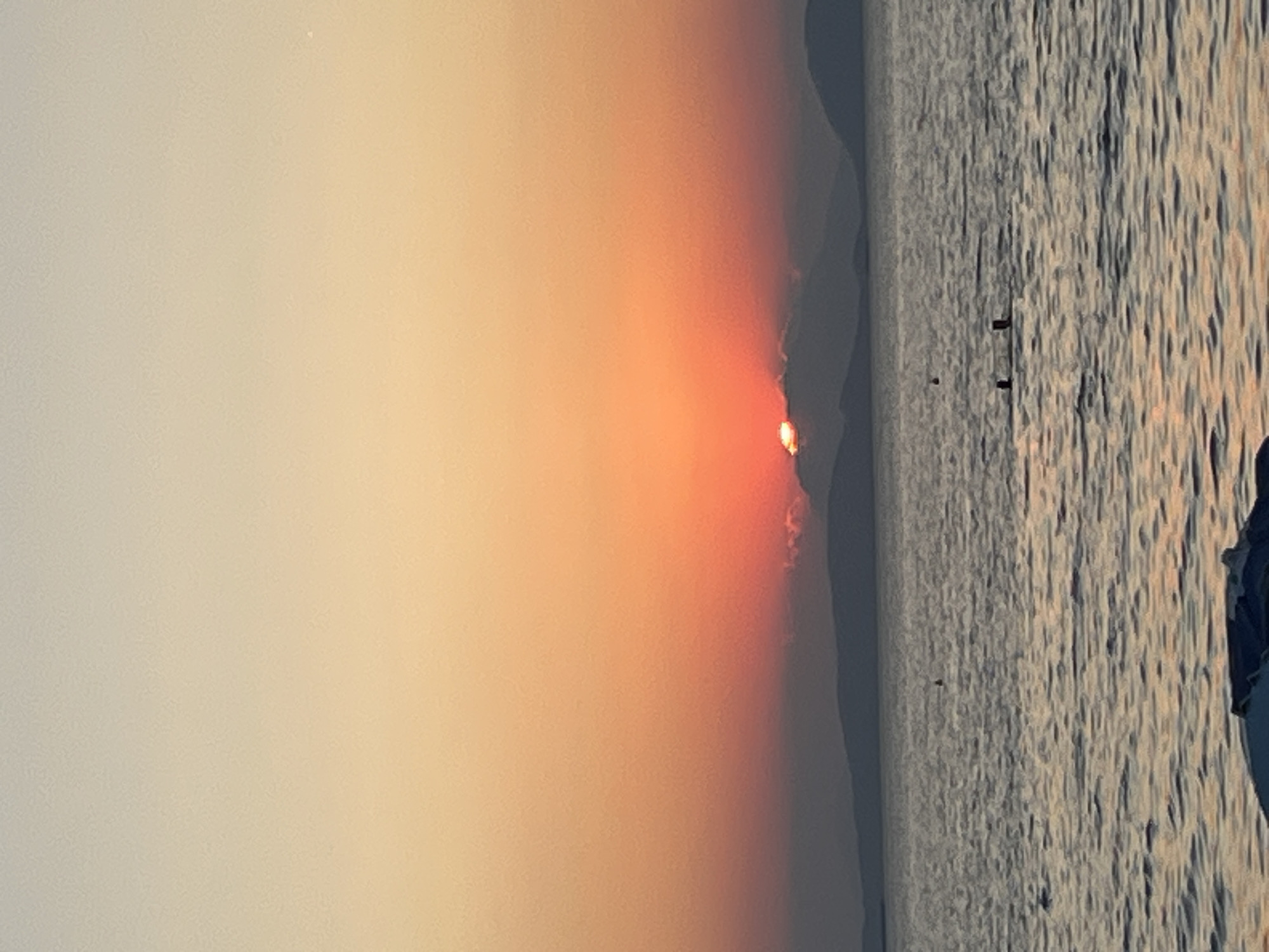 The image captures a serene sunset over the Aegean Sea, likely near Vouliagmeni Municipal Unit, Greece, given the coastal landscape and the typical sunset colors. The sky transitions from a pale yellow at the top to a vibrant orange and red near the horizon. Silhouetted mountains form a gentle curve across the distance, with the sun partially obscured by wispy clouds just above them. The sea is calm, reflecting the warm hues of the sky, with small ripples on its surface. In the middle ground of the water, there are several small, dark objects, possibly buoys or markers. Two figures are visible in the water, appearing to be swimming or wading. In the foreground, partially cropped from the bottom of the frame, is a section of a boat or inflatable raft, suggesting a location near a beach or harbor. The overall atmosphere is tranquil and picturesque, characteristic of a Mediterranean sunset.