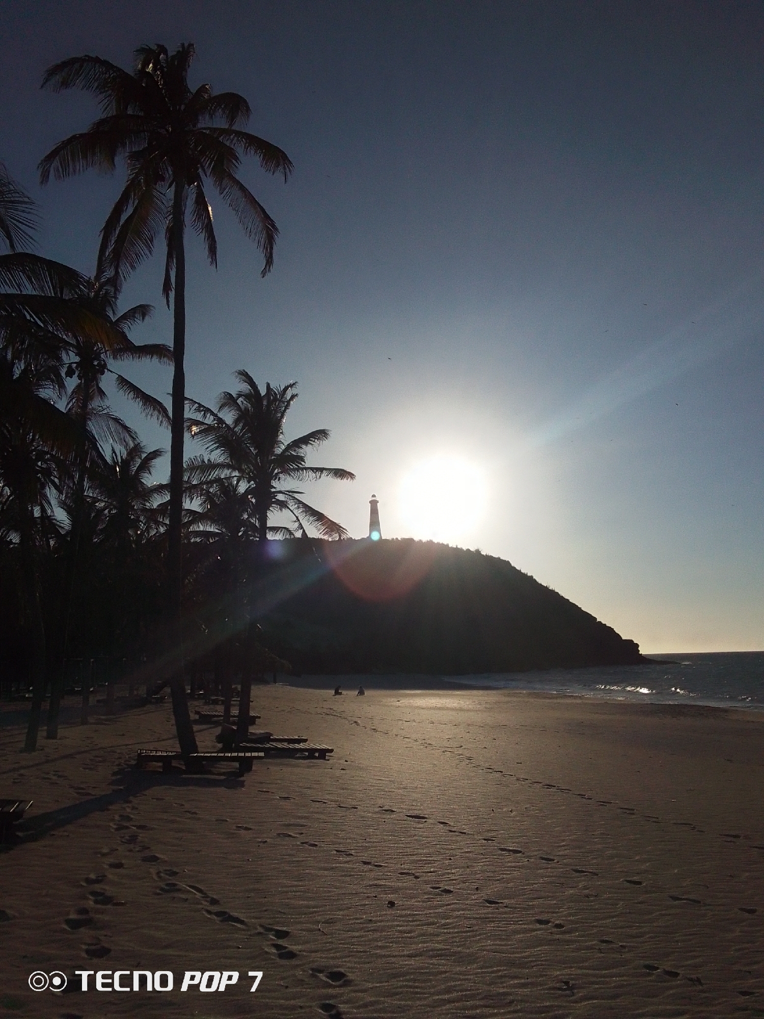 The image depicts a beach scene in Pedro González, Venezuela, at sunset. The sun is positioned over a small hill, silhouetting a lighthouse structure. A row of palm trees lines the left side of the image, casting shadows across the sandy beach. Several lounge chairs are visible beneath the trees. Footprints are scattered across the beach leading towards the water. The ocean stretches along the right edge of the scene. The text "TECNO POP 7" is visible in the lower left corner of the image.